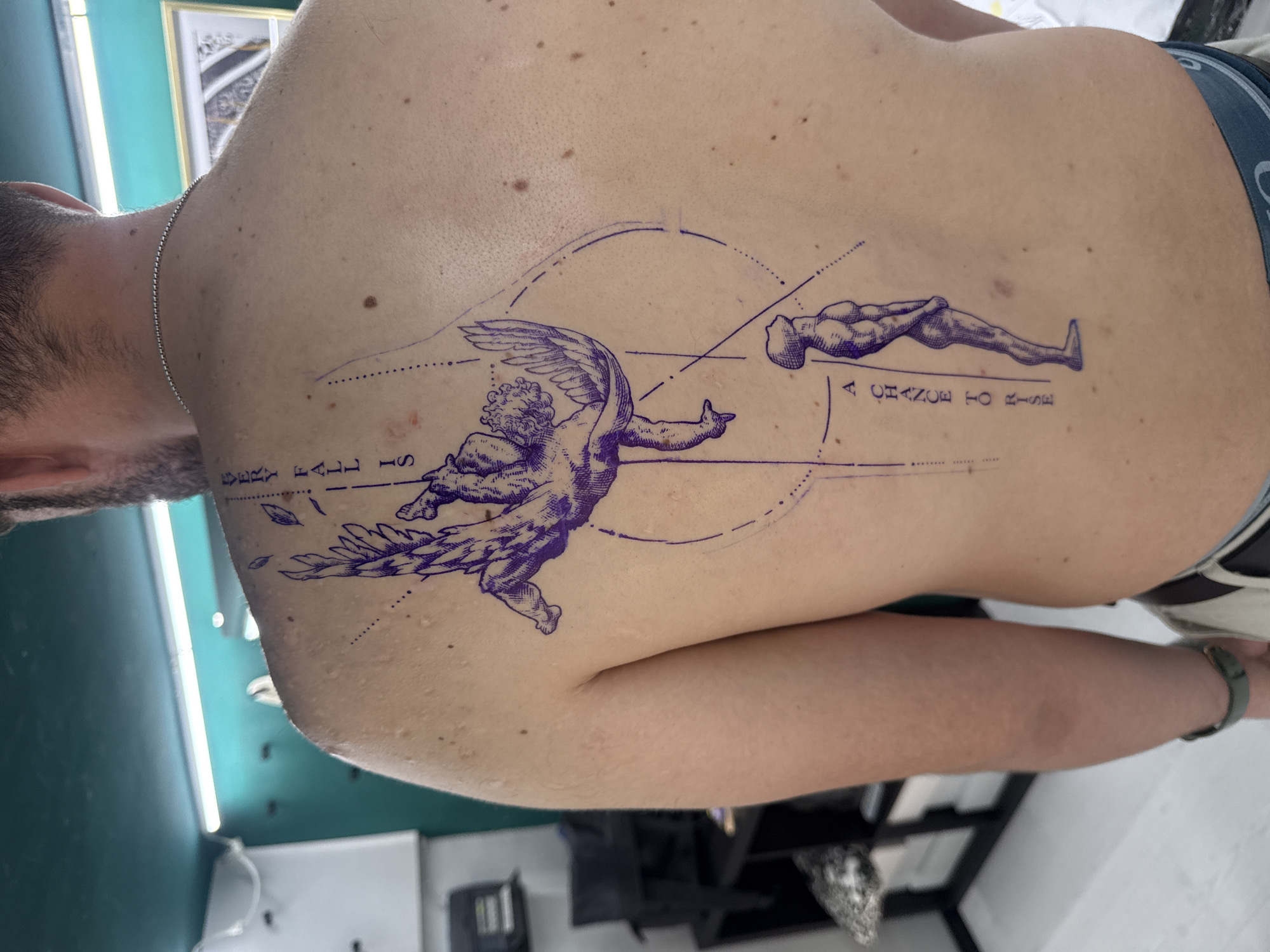 A close-up, medium-shot photograph shows a person's back with a fresh tattoo. The tattoo, rendered in dark blue ink, features an angel-like figure with wings in a dynamic pose, along with a prone, muscular figure lying horizontally above it. The phrase "A CHANCE TO RISE" is tattooed next to the prone figure, and the phrase "EVERY FALL IS" is tattooed next to the winged figure. The tattoo is executed with fine lines and shading. The person in the photo has fair skin with some freckles. The background reveals a room with a green wall and a glimpse of what appears to be a tattoo parlor or similar setting. The location is not discernible from the image.