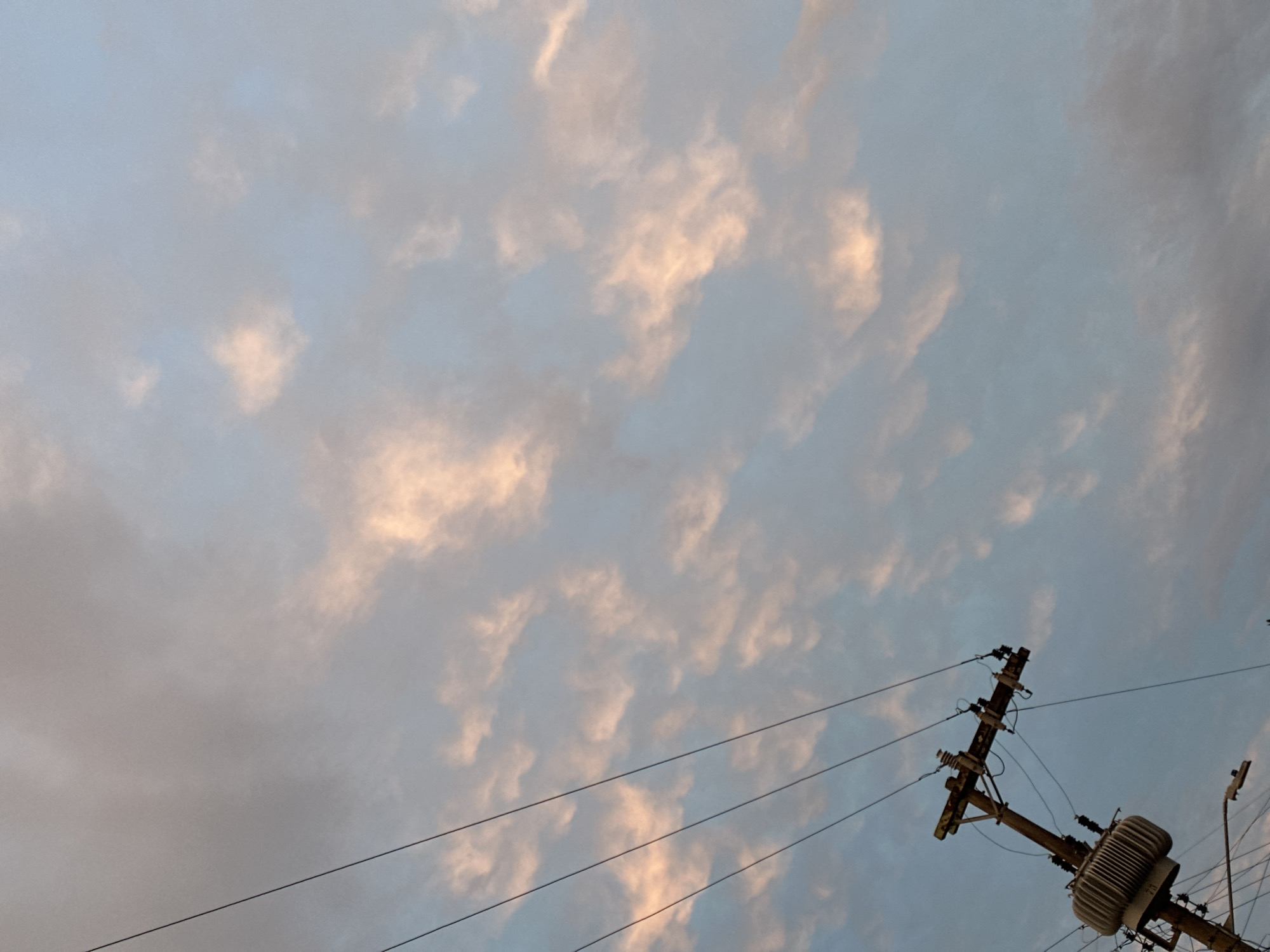 The image shows a low-angle view of the sky, filled with scattered, mostly white clouds tinged with light orange. At the bottom right corner, there is a wooden utility pole with electrical wires and a transformer. The sky appears to be mostly clear, with a mix of light blue and grey tones. The photograph was likely taken in an urban area due to the presence of the utility pole, but without further information, the specific location in Florianópolis, Brazil, cannot be determined. There are no people visible in the image.