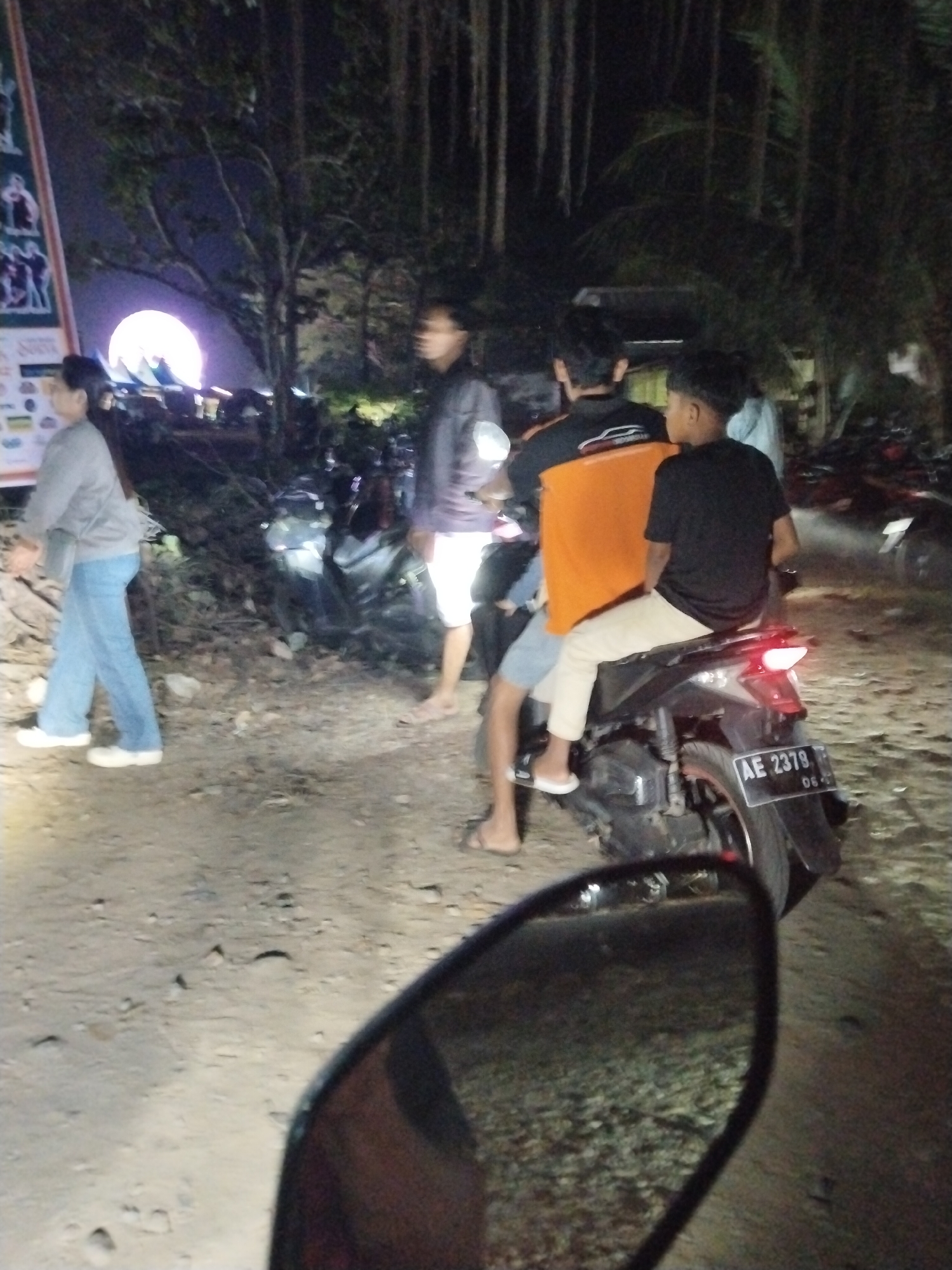 A nighttime outdoor scene shows several individuals and a motorcycle. In the foreground, a person is reflected in a motorcycle's side mirror. To the left, a woman in a gray top and blue jeans walks past a sign with graphics. Behind her, a large, brightly lit, circular object, possibly an inflatable moon, is visible, illuminated by unseen lights, with trees and structures in the background. Further back, three young men are gathered around a motorcycle. Two are seated on the motorcycle, one in an orange shirt and the other in a black shirt. The man in the orange shirt has his arm around the man in the black shirt. The third young man stands nearby. The motorcycle has a license plate reading "AE 2379". The ground is dirt and gravel, and there are some scattered rocks. The overall environment suggests a public gathering or event in a dimly lit area, possibly near a road or parking lot in Tasikmadu, Indonesia.
