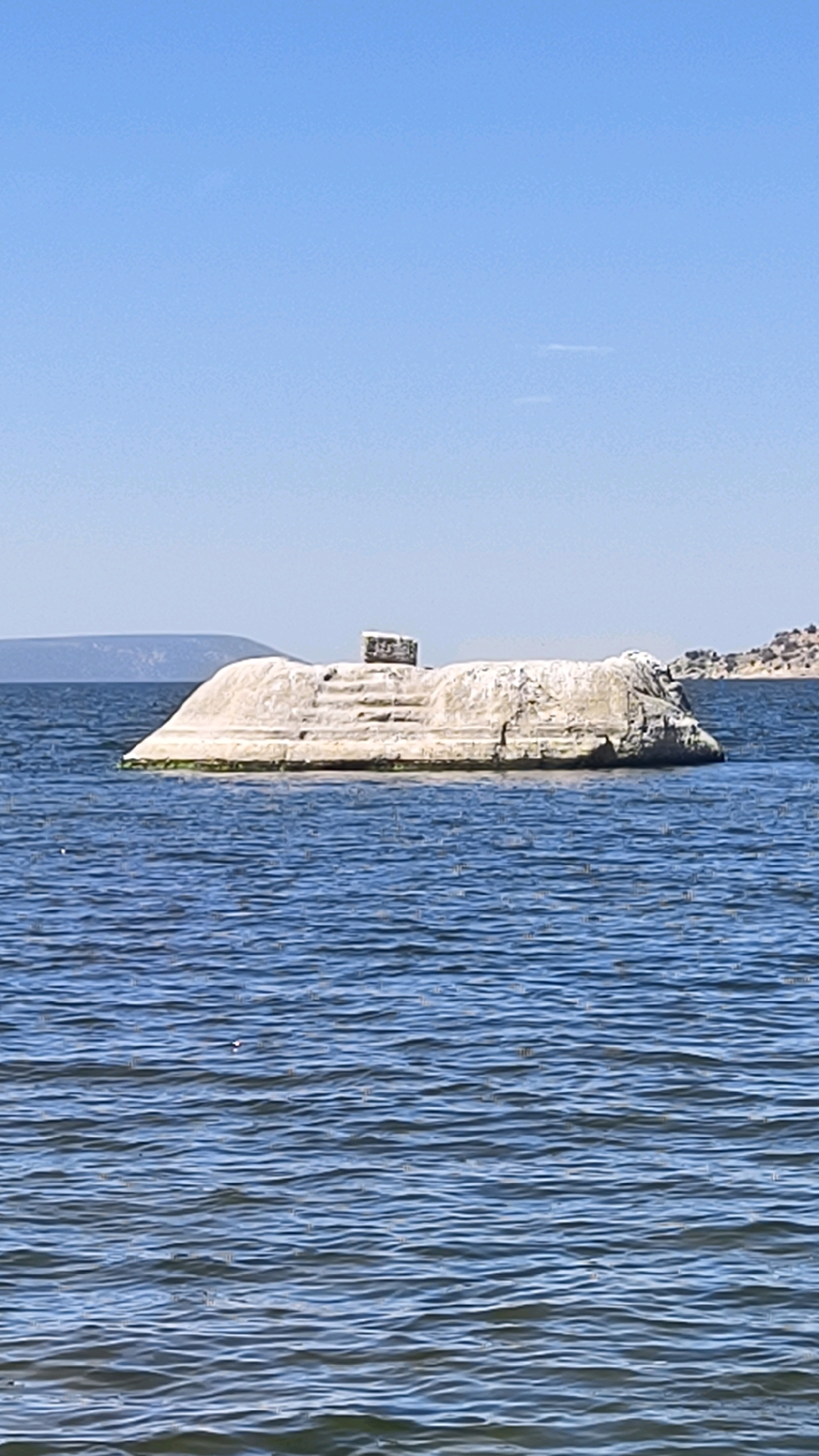 A large, weathered rock formation, appearing to be a naturally occurring island or a man-made structure partially submerged, sits in the middle of a body of blue water under a clear sky. The rock is light-colored, with visible texture and some green growth near its base. On top of the rock, there are what appear to be carved steps leading up to a square, block-like structure. The water is choppy with small waves. In the distance, a hazy shoreline with hills can be seen, and on the far right, more rocky terrain is visible. The overall scene suggests a natural landscape, possibly a lake or a calm sea.