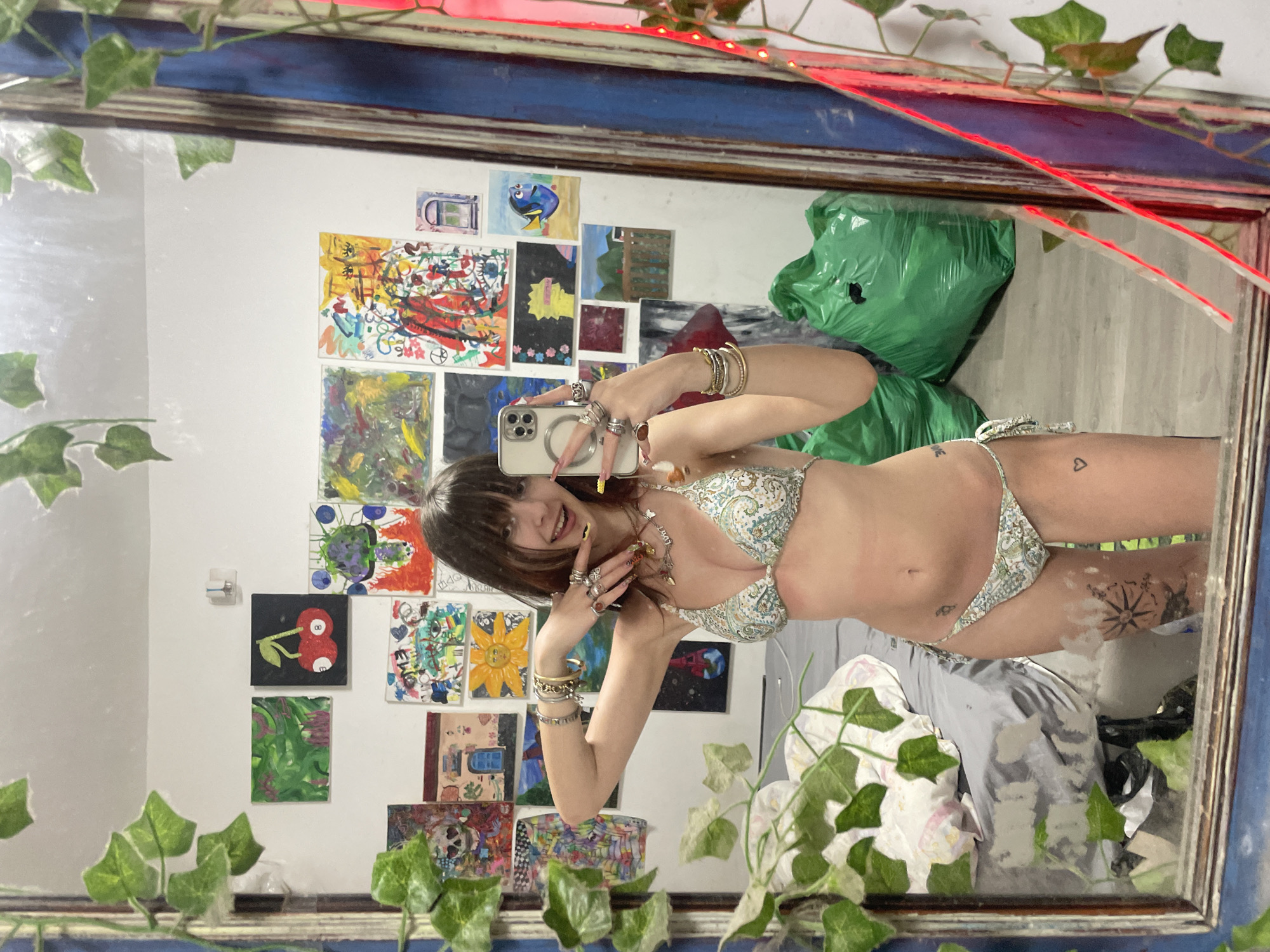 A young woman is taking a selfie in a mirror. She is wearing a bikini and jewelry on her arms and fingers. Her hair is brown with blonde highlights. The background consists of a white wall with a variety of small paintings and a green trash bag. The mirror frame is decorated with artificial ivy and red lighting. The image does not indicate the location in Raanana, Israel.