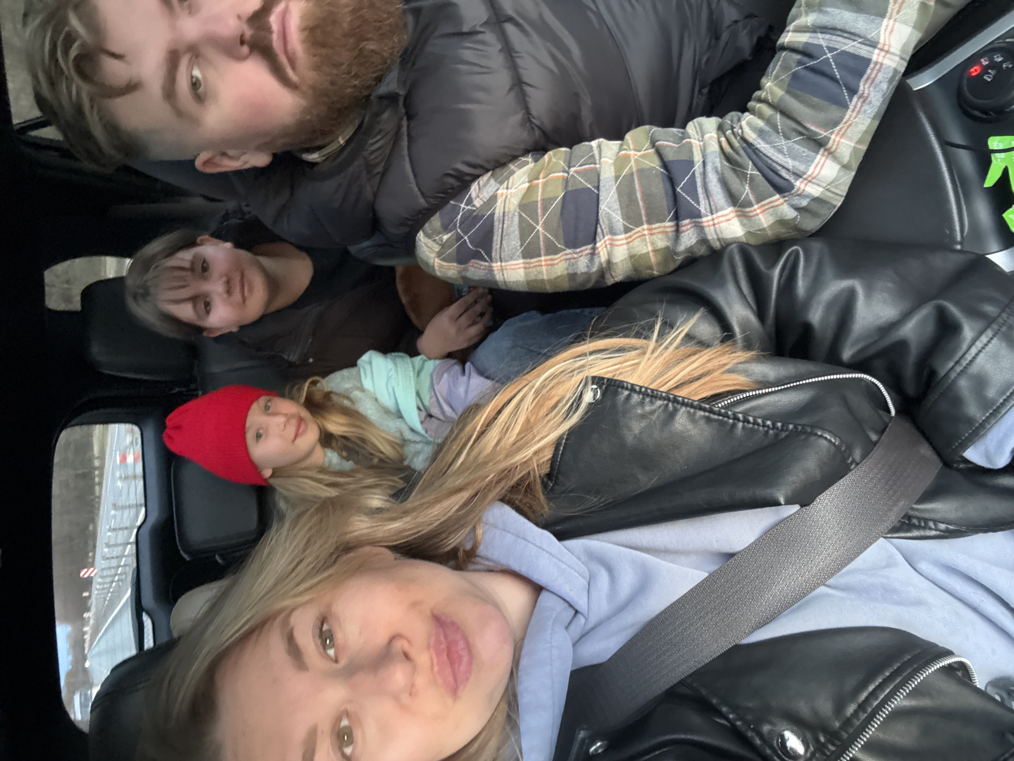 Four people are in a vehicle. From the perspective of the camera: a blonde woman in a black leather jacket and blue hooded shirt is in the front; a younger woman in a red knit cap is behind her; further back is a person with short dark hair. In the far back corner is a bearded man in a dark puffer coat and plaid shirt. The camera is likely held by the woman in the front. The location appears to be near a road, possibly a highway, in Gubarevo, Russia. There is no definitive evidence to confirm the specific location as Gubarevo, Russia.
