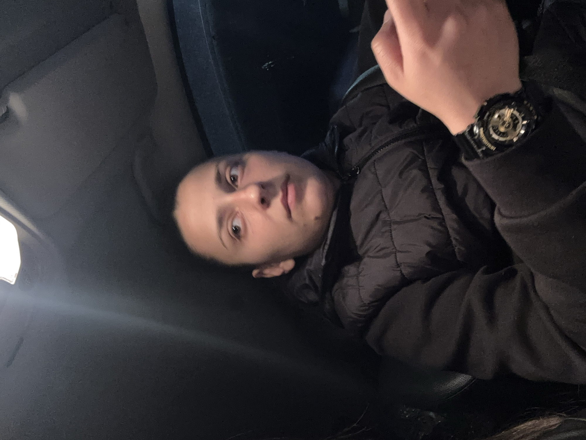 A person with short dark hair and light skin is seated in a car. The person is wearing a black padded jacket and a black watch on their left wrist. The interior of the car is dark, with the dashboard and a portion of the car's ceiling visible. A light source is visible in the upper left corner of the image, likely a streetlight or the car's interior light. The location in Sofia, Bulgaria cannot be determined from the image.