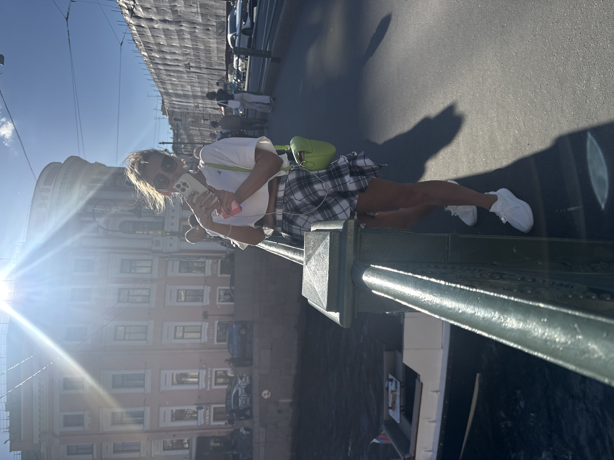 A woman with blonde hair pulled back stands on a bridge in Saint Petersburg, Russia, holding a smartphone to take a selfie. She is wearing sunglasses, a white cropped t-shirt, and a black and white plaid mini-skirt with a ruffled hem. A bright green crossbody bag is slung over her right shoulder. She is wearing white sneakers. The background shows a canal with a boat on the left, and buildings with ornate architecture and construction scaffolding on the right. The sun is shining brightly, creating lens flare and casting long shadows on the asphalt bridge. Other pedestrians are visible in the background.