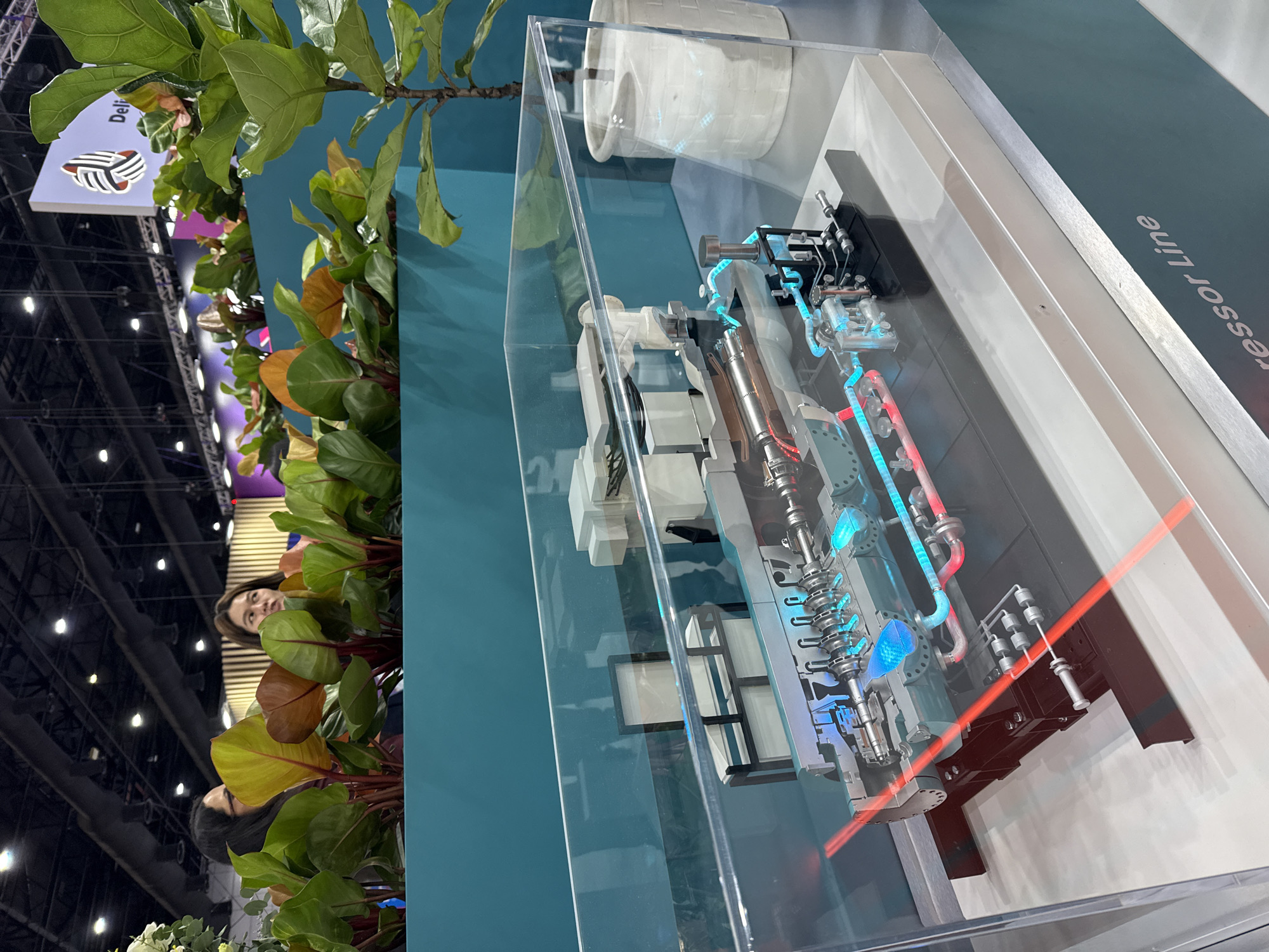 An indoor event space, likely a convention or exhibition hall, features a detailed model of a compressor unit displayed under a clear acrylic casing. The model showcases a cross-section of the compressor, highlighting its internal workings with illuminated blue and red elements representing fluid flow. The surrounding environment includes lush green plants in teal planters, creating a vibrant contrast with the industrial exhibit. In the background, blurred figures of two women can be seen, one wearing glasses. A partial view of a sign with "Deli" and a stylized logo is visible in the upper right corner. The scene appears to be set up for a trade show or product demonstration. The floor is a neutral grey. The image does not provide any direct information about the city of Bangkok, Thailand.