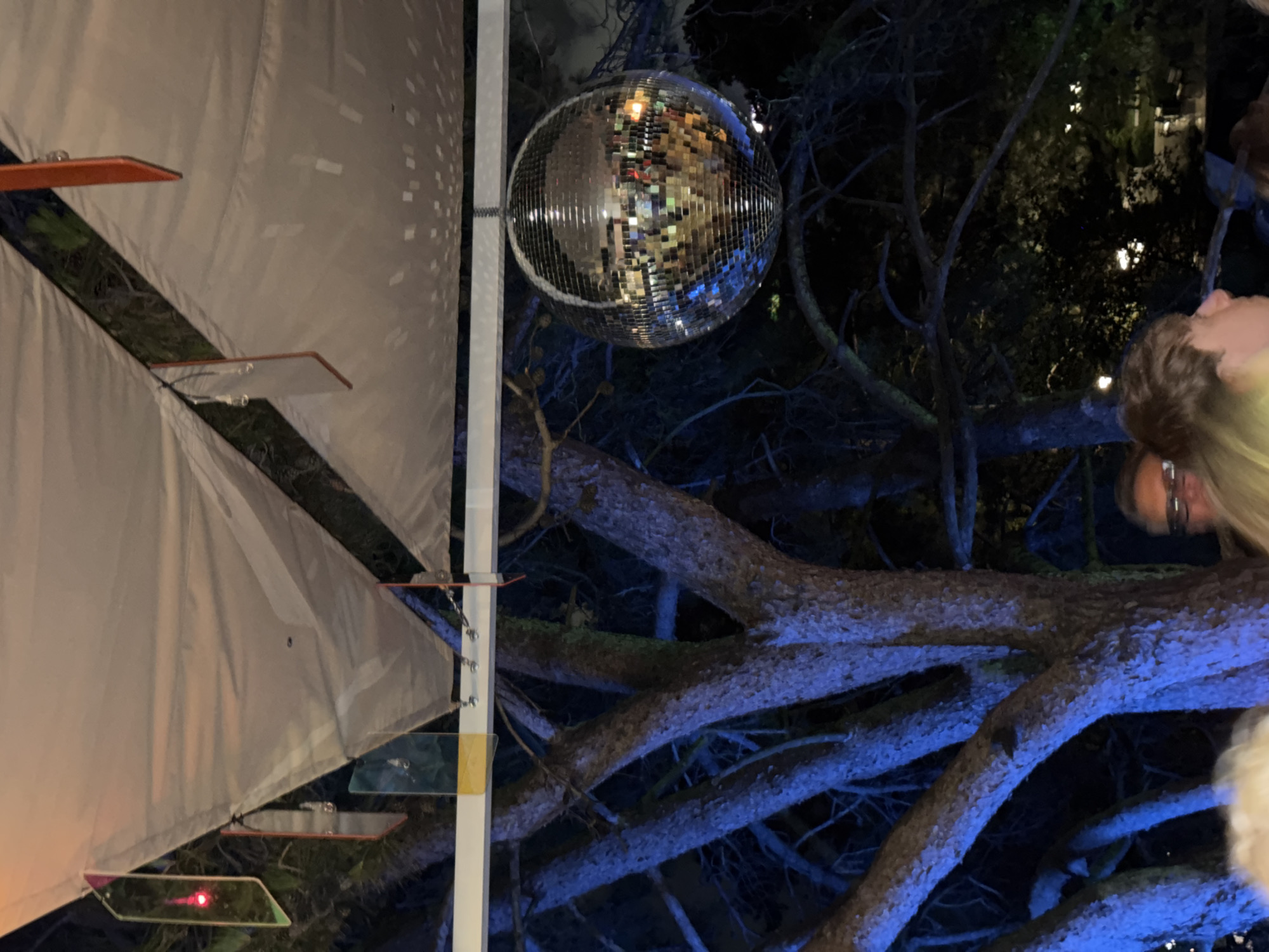 This is a medium shot, outdoor photograph taken at night, featuring a disco ball hanging from a white canopy structure. The disco ball is centrally located and reflects ambient lights. To the left of the disco ball, a tree with rough bark is illuminated with blue light, its branches extending into the darkness. Several rectangular, colored translucent decorations, possibly made of acrylic, are suspended from the canopy. Some of these decorations show reflections or colored lights, with one on the far left displaying a red light. The background is dark with hints of foliage and some distant lights, suggesting an outdoor venue. The lower part of the image shows the heads and shoulders of several individuals, appearing to be patrons of an event, with some illuminated by a yellow-green light. The context provided about Sochi, Russia, is not discernible from the image itself.