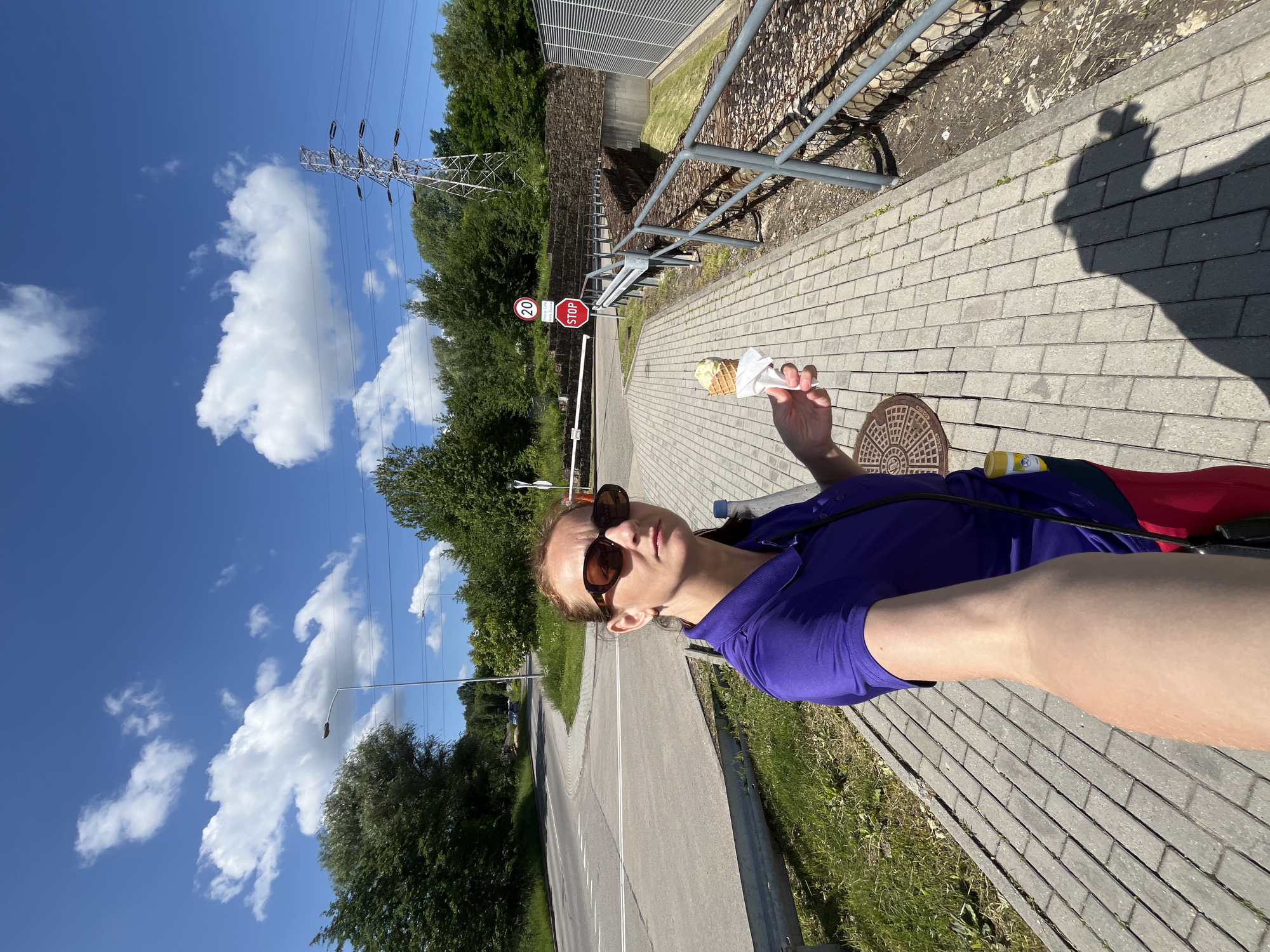 A woman stands on a paved sidewalk, holding an ice cream cone in her right hand. She is wearing a purple polo shirt, sunglasses, and red pants. The woman is taking a selfie, as indicated by her arm extended towards the camera. In the background, there is a road with white lane markings, trees, a speed limit sign with "20" on it, a red STOP sign, a metal barrier, and a large power line tower. The sky is blue with white clouds. The scene appears to be outdoors in a semi-urban or rural setting, possibly near a road junction or entrance.