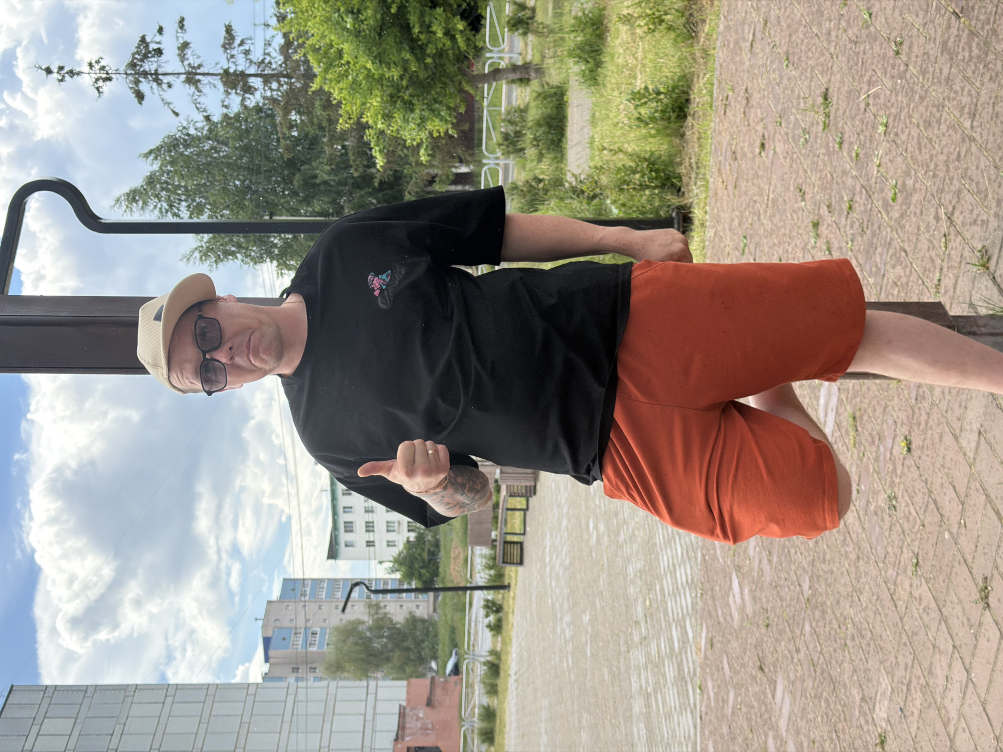 A man wearing a black t-shirt, orange shorts, a light-colored hat, and glasses stands outside in front of a pole. He is giving a thumbs-up gesture. In the background, there are apartment buildings, trees, and a cloudy sky. The ground is paved with bricks. The location is the city of Chaikovsky, Russia.