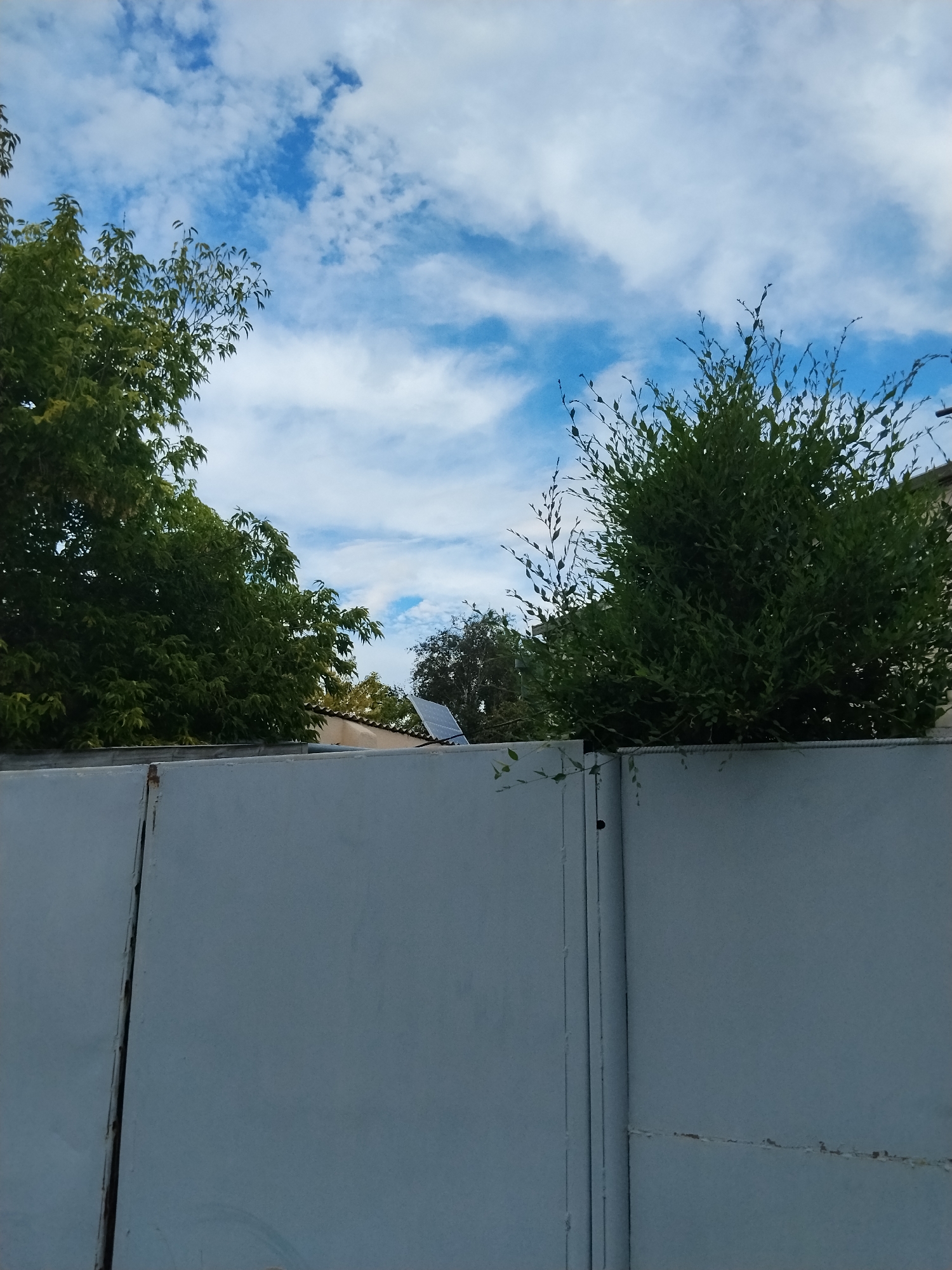 The image shows a gray metal gate in the foreground, with trees and a blue sky with white clouds in the background. A solar panel is visible on a roof behind the gate. The scene appears to be a residential area in Kokshetau, Kazakhstan. There are no people or animals visible in the image. The lighting suggests it is daytime.