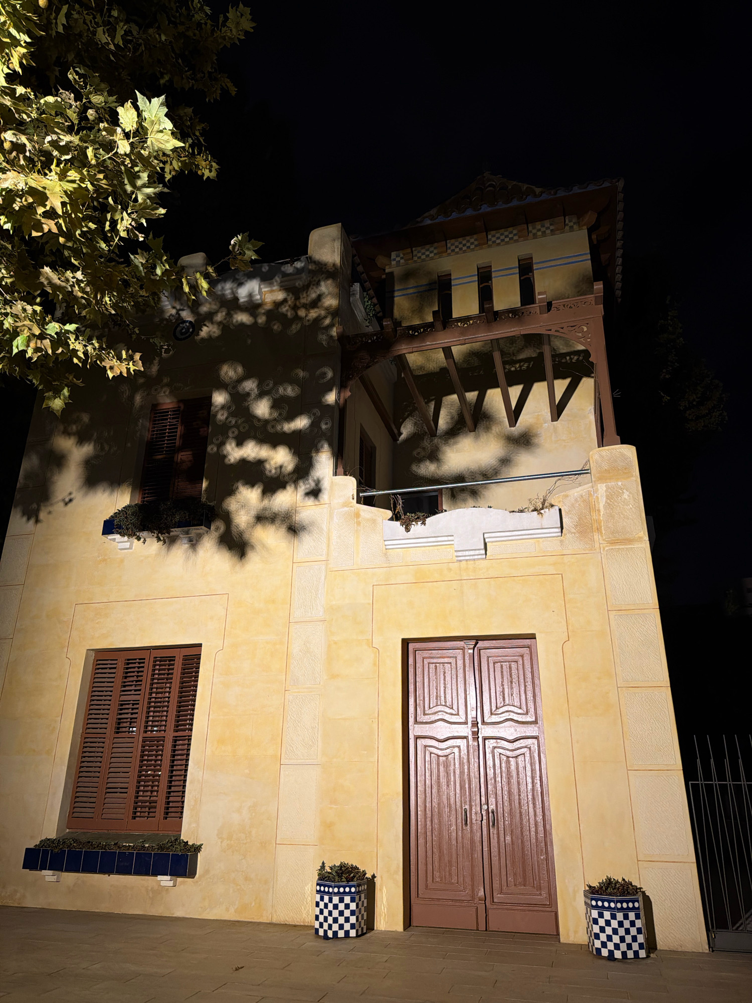 The image shows a close-up, low-angle shot of a tan-colored building at night. The building features a dark brown, louvered window on the left side, with a planter box below it. To the right of the window, there is a decorative cornice and a balcony with a railing. The main entrance to the building is a set of double doors, also dark brown, with carved panels. Two blue and white checkered planters, each containing a small plant, are positioned on the paved ground in front of the building. Above the main section of the building, there is an ornate, multi-tiered roof structure with visible wooden beams and decorative tiles. A large tree with yellowing leaves is visible in the upper left corner, casting dappled shadows on the building's facade. The background is dark, indicating nighttime, with a hint of dark foliage to the right.
This building is identified as a part of the historic Cap de Salou architecture in Salou, Spain, known for its distinctive style and placement along the coastline.