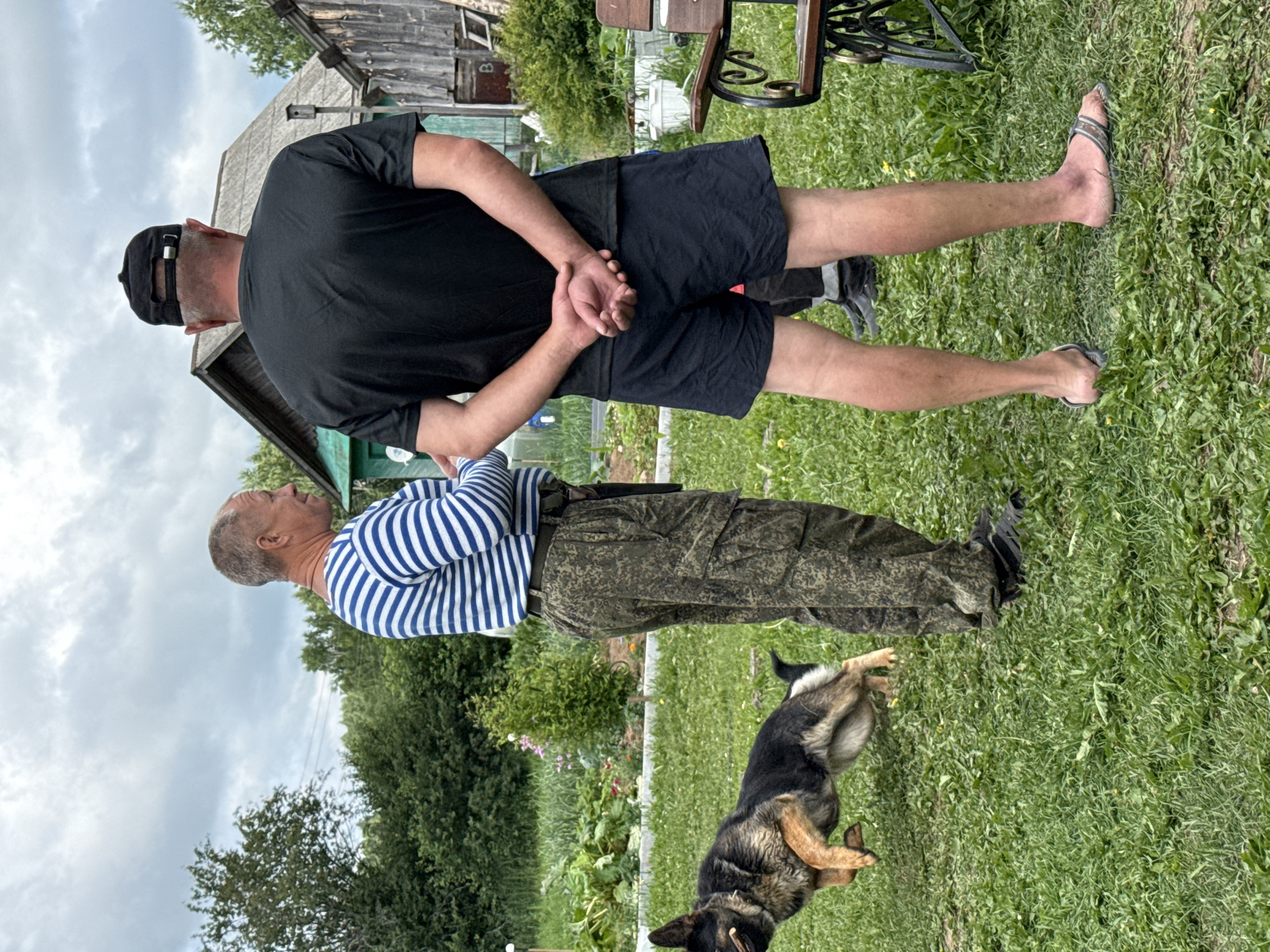 An outdoor scene in what appears to be a rural area of Unknown, Russia, depicts two men and a German Shepherd dog. The man on the left is wearing a blue and white striped long-sleeved shirt and camouflage pants. He is facing to the left, with his body angled towards the dog. The dog, a German Shepherd, is mid-air in a jump, facing left, with its mouth open and a stick or chew toy in its mouth. It is positioned to the left and slightly in front of the man in the striped shirt. The man on the right is wearing a black t-shirt, black shorts, a black baseball cap, and flip-flops. He is facing away from the camera, with his hands clasped behind his back. He is standing to the right of the other man. The environment includes a grassy yard with scattered yellow wildflowers. In the background, there are trees, a green building, and wooden structures suggesting a rustic residential area. A portion of a dark metal bench with ornate scrollwork is visible in the lower right corner of the image. The sky is overcast with a cloudy, gray appearance.