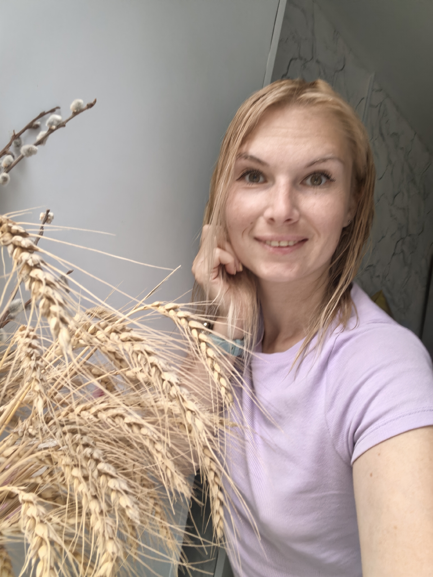 A blonde woman with light brown eyes smiles at the camera, leaning her chin on her hand. She is wearing a light purple t-shirt and has a blue watch on her left wrist. In the foreground, a bouquet of dried wheat stalks and pussy willow branches are arranged in front of her. The background appears to be an indoor setting with a light gray wall and a textured gray surface to the right. The image has been taken from a selfie perspective.