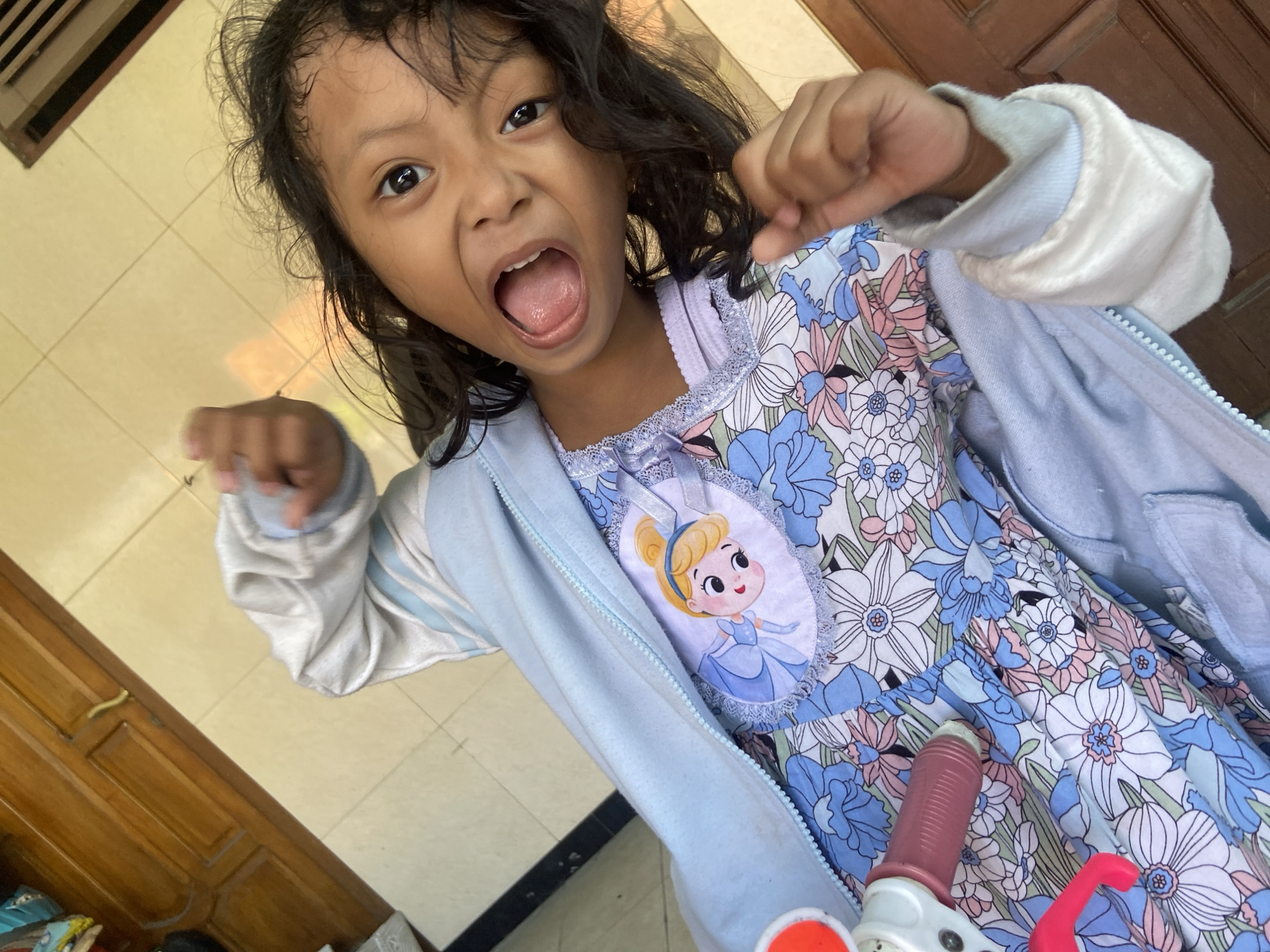 A young girl with dark hair and open mouth is shown from a high angle, making a playful gesture. She is wearing a blue zip-up jacket over a floral print dress featuring pink and white flowers. A white bib with a cartoon depiction of Cinderella is attached to the dress. A toy, possibly a part of a stroller or walker with a red handle, is visible on the left side of the frame. The background consists of light-colored tiled flooring and a wooden door. The scene appears to be outdoors, possibly in a residential area of Gelam, Indonesia, given the architectural elements and flooring.