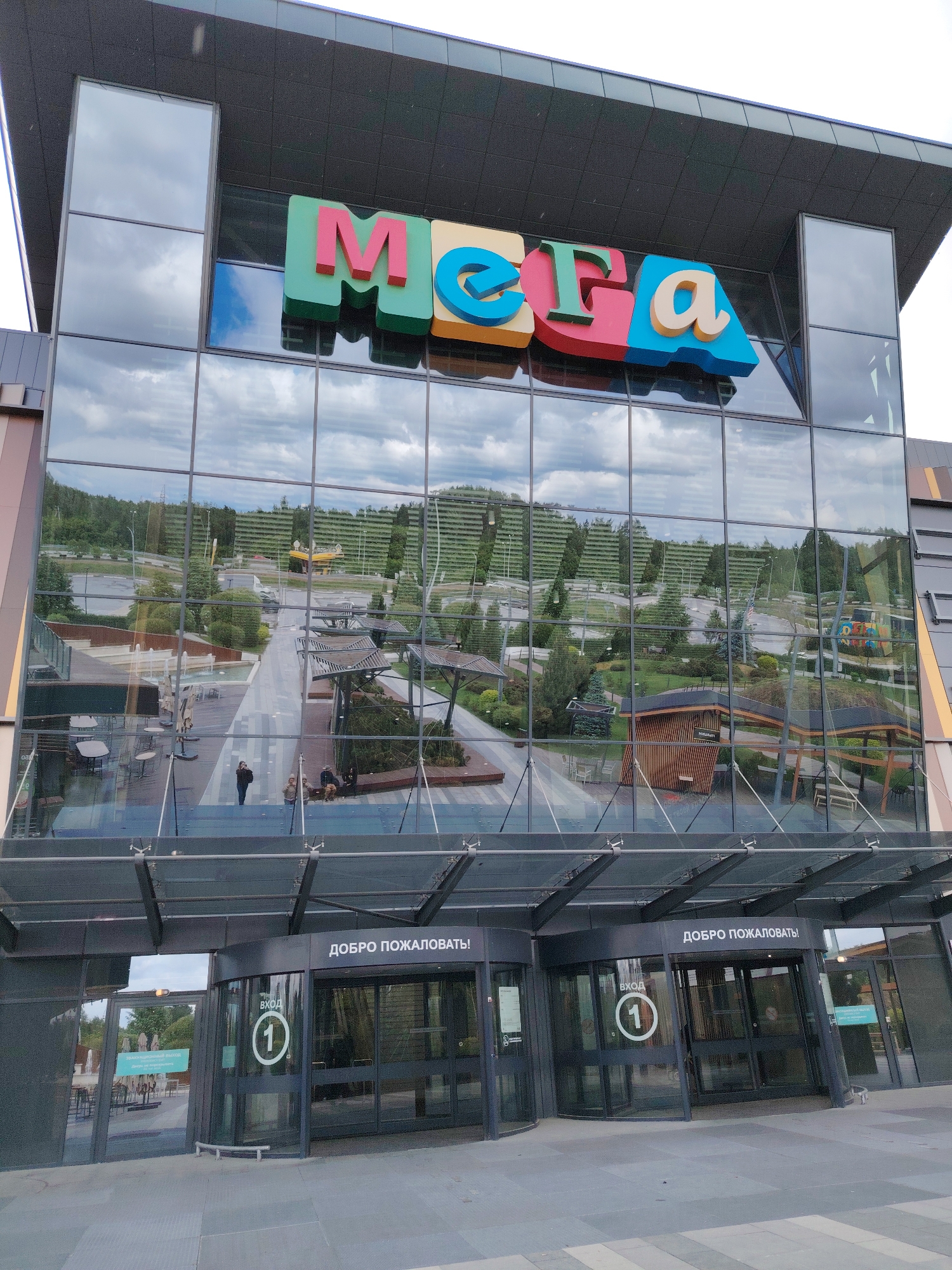 The image shows the facade of the MEGA shopping center in Yekaterinburg, Russia. The building has a large glass exterior that reflects the surrounding landscape. Above the entrance is the large, colorful MEGA logo. The entrance features revolving doors with signs in Russian that say "Welcome" and "Entrance 1". A few people are visible near the entrance in the reflection of the glass windows. The reflection suggests there is a courtyard or outdoor area with seating and landscaping features.