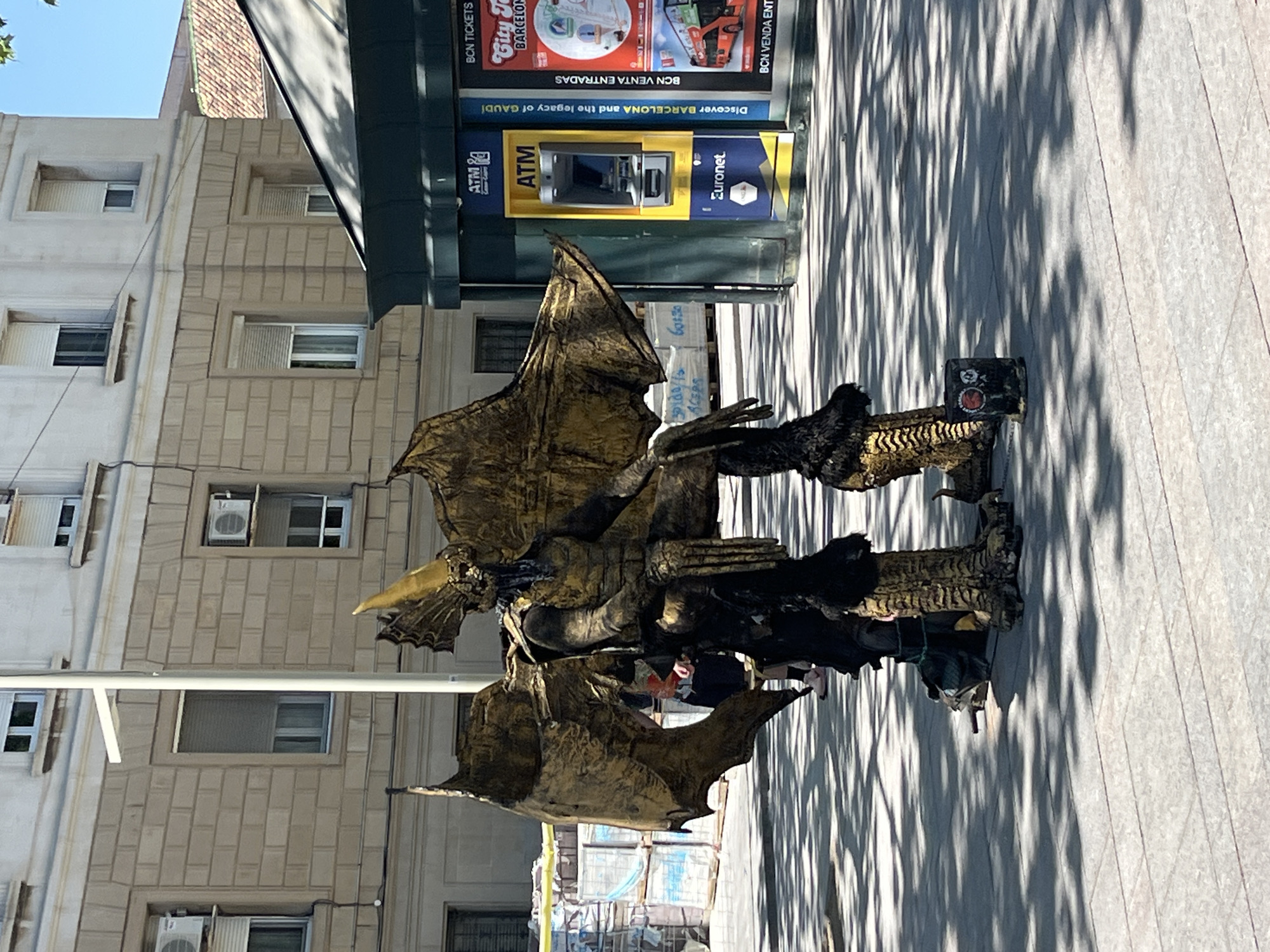 Here's a detailed description of the image:
The photograph shows a street performer dressed in an elaborate golden dragon costume. The costume includes large wings and a detailed head with a beak. The performer is standing on a paved area, presumably a pedestrian zone. Behind the performer is a building with multiple windows and air conditioning units.
Next to the performer is an ATM machine with advertisements for Barcelona attractions. The advertisement includes mentions of "Discover Barcelona and the legacy of Gaudí" and a promotion for "BCN Tickets, City Tours Barcelona." The ATM also displays "ATM Euronet."
Shadows from nearby trees or structures fall across the pavement, creating a pattern of light and dark. The environment suggests the photo was taken during daylight in Barcelona, Spain.