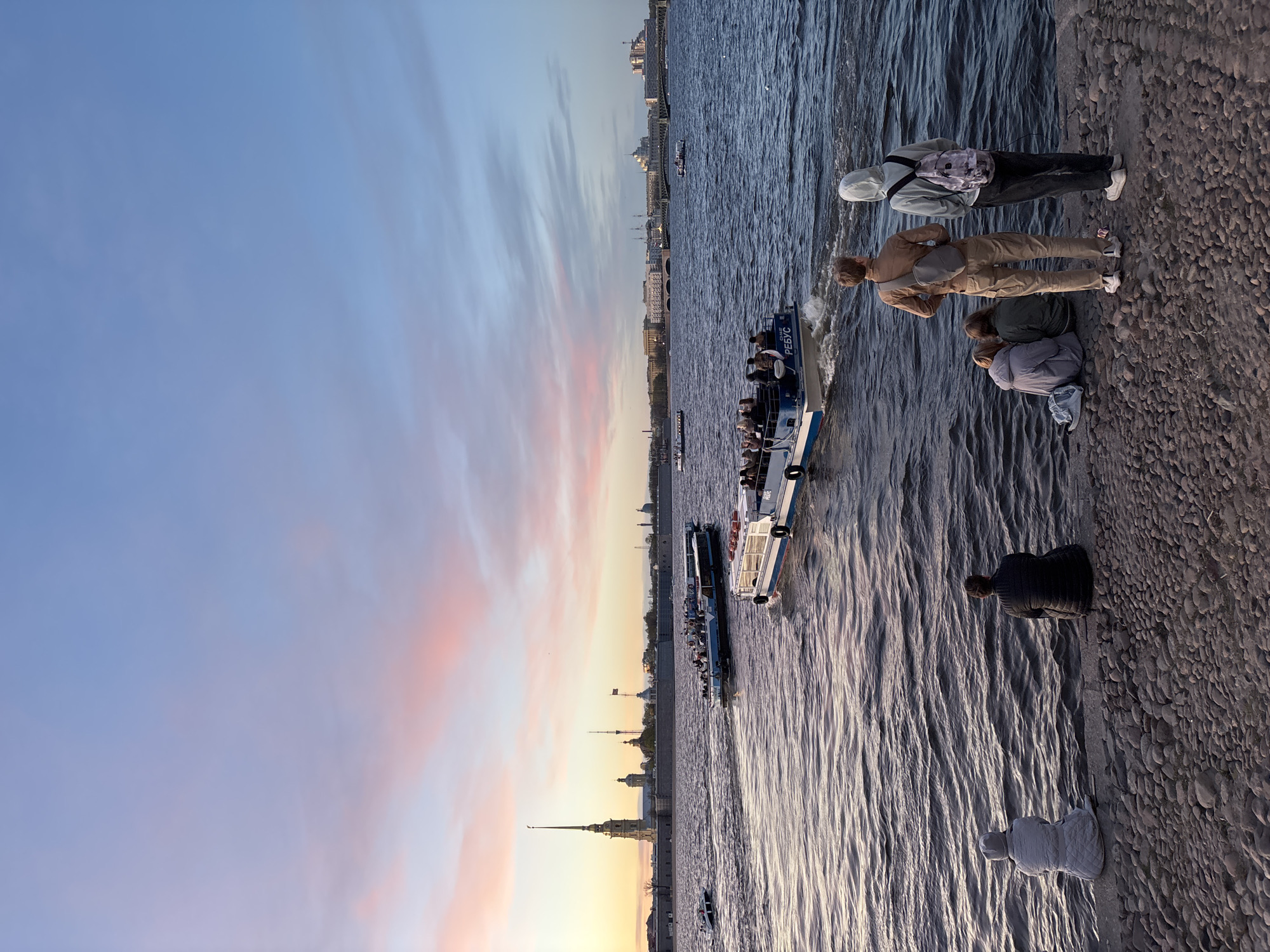 The image shows a view of Saint Petersburg, Russia, along the Neva River at sunset. The sky displays a gradient of blue and pink hues. Several tour boats filled with people traverse the river. On the riverbank, people are observed; some are looking towards the water while others are walking. Landmarks of Saint Petersburg, including the Peter and Paul Fortress, are visible in the background skyline. The foreground consists of the stone embankment and the water's surface, marked by ripples.