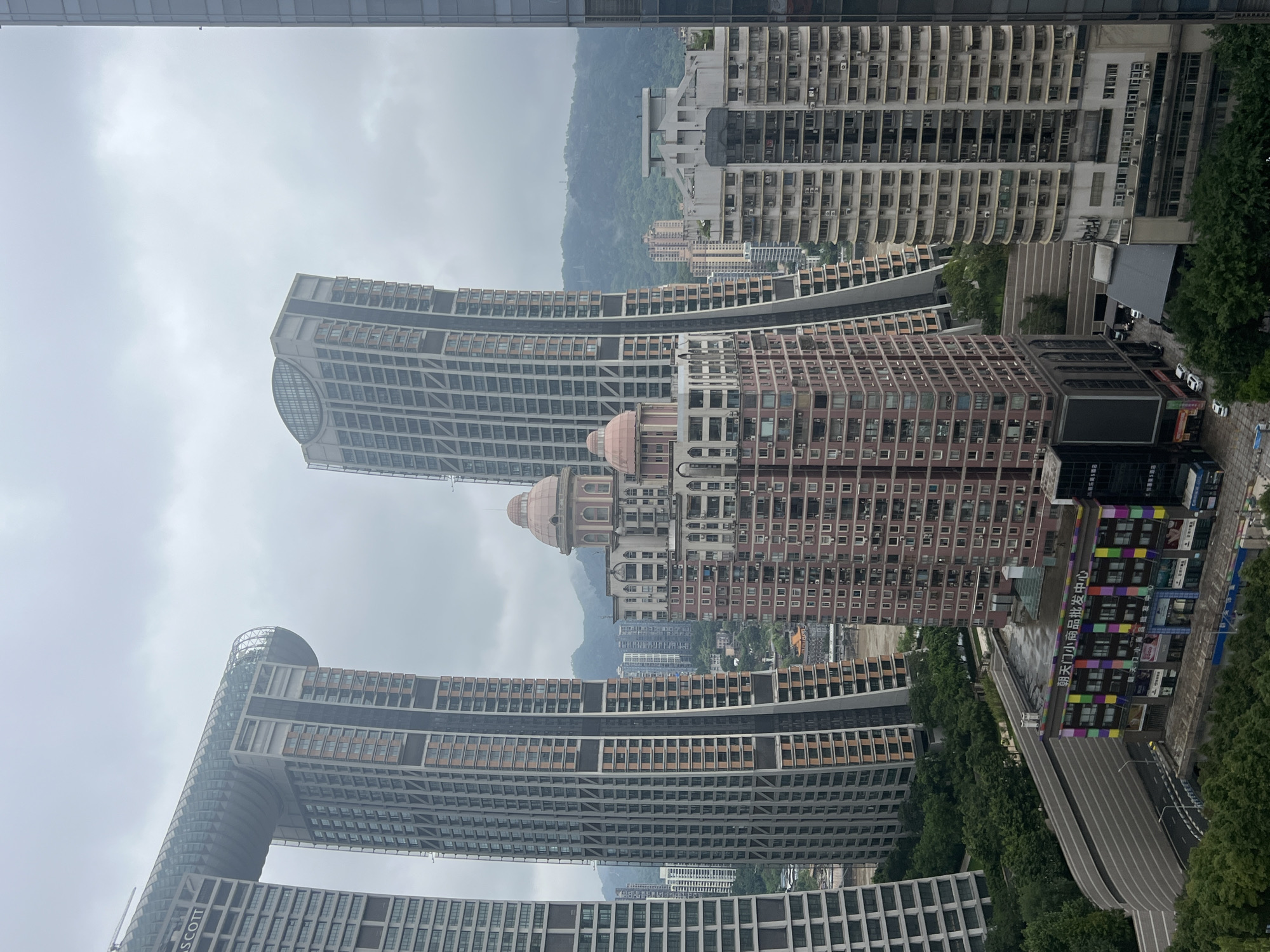 This is an aerial view of a dense urban landscape in Yuzhong District, Chongqing, China. The scene is dominated by a collection of contemporary high-rise buildings with varied architectural styles. Prominently featured is a unique structure consisting of two towers connected by a horizontal section at their upper levels, creating a large archway. This building has a curved, glass-enclosed rooftop garden. To its right, another tall, slender skyscraper with a curved facade rises into the overcast sky. In the foreground and middle ground, several other residential and commercial buildings are visible, including a pink building with dome-like structures on its roof, and a lower, colorful building with Chinese signage that appears to be a wholesale market. The background reveals rolling green hills under a cloudy sky. Trees and greenery are interspersed between the buildings, suggesting a city with a significant amount of vegetation. The image captures a bustling, modern city environment.
