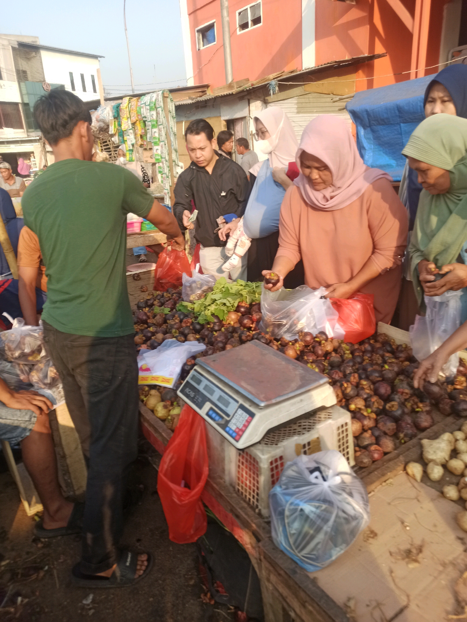 The image depicts a bustling outdoor market scene, likely in Cikarang, Indonesia, with a focus on the sale of fresh produce. In the foreground, a vendor, a man wearing a green t-shirt and dark pants, is interacting with a customer. He is standing behind a wooden stall laden with mangosteens, a common fruit in Southeast Asia. Several customers are visible, including women wearing hijabs, who are examining the fruit. One woman in a peach-colored hijab is holding a mangosteen in her hand, possibly inspecting it. A digital scale sits on the counter, ready for weighing purchases. In the background, there are stalls selling various items, including packaged goods and other produce. Buildings with colorful facades, typical of urban Indonesian architecture, can be seen in the distance. The overall atmosphere is one of lively commerce, with people engaged in the daily activities of a local market.