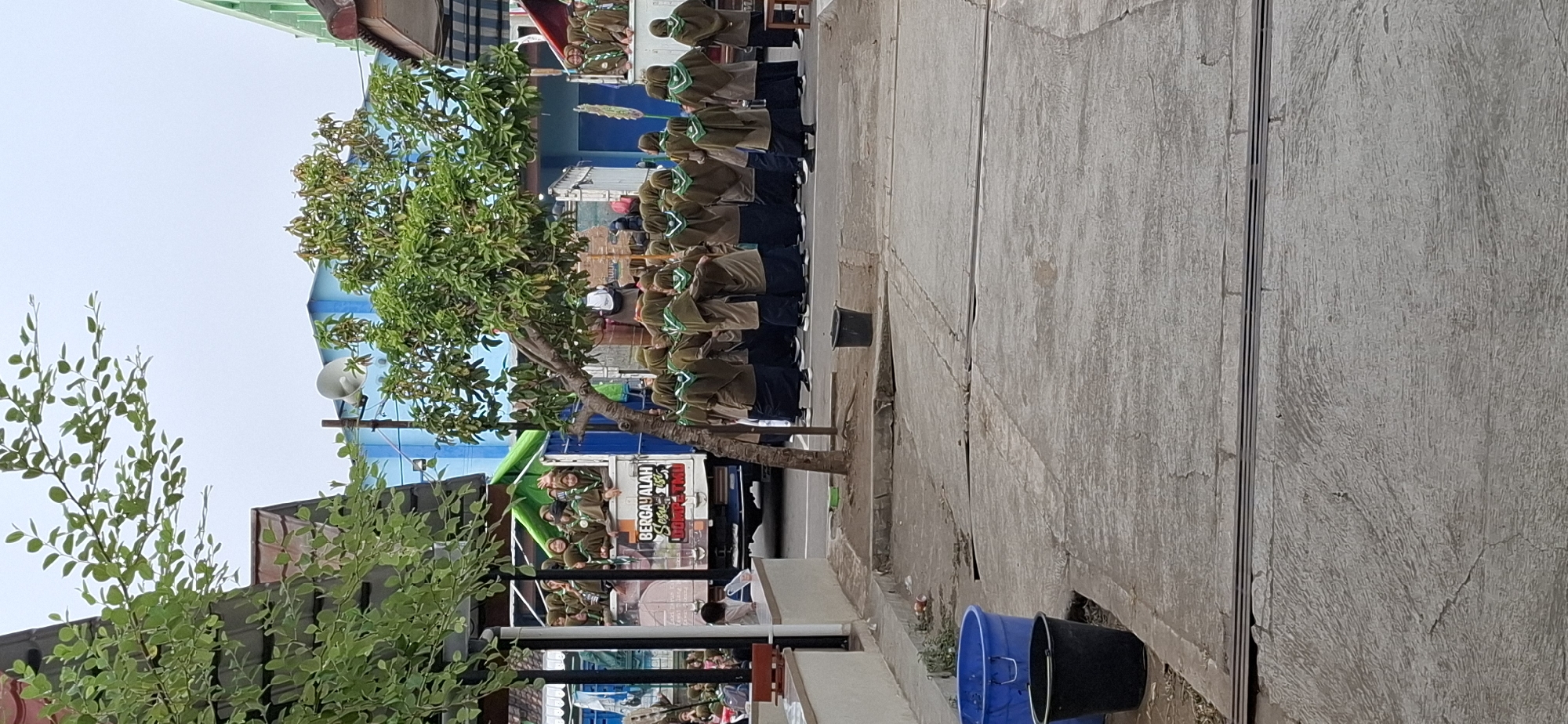 In Wonorejo, Indonesia, a group of young people in matching olive green uniforms and headscarves are gathered near a truck with a green tarp. Some are standing on the truck bed, waving, while others are clustered on the ground in front of it. A large tree with green leaves stands in the center of the frame, partially obscuring the scene. In the background, a blue building with white trim is visible, along with a white loudspeaker mounted on a pole. Two large plastic bins, one blue and one black, are in the foreground on a concrete surface, with debris scattered around them. The overall environment appears to be an outdoor public space, possibly near a school or community center.