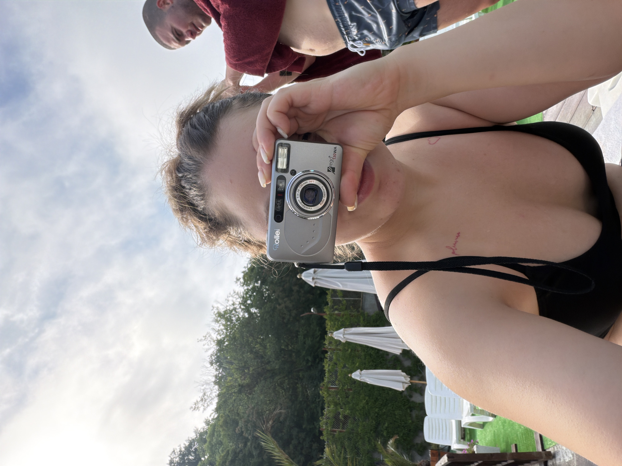 The image is an outdoor, low-angle shot, presented as if rotated 90 degrees clockwise. The primary subject is a person, likely female, holding a silver Rollei compact digital camera directly in front of their face, obscuring their eyes. The person is wearing a black bikini top, and a small red heart tattoo is visible on their upper chest, with another faint red script tattoo (possibly "pluma") on their right collarbone area.
In the upper background, another person, likely male, is visible upside down due to the camera's orientation. They are wearing a red top and dark patterned shorts. The sky is bright with scattered white clouds, indicating a sunny daytime setting.
The camera held by the main subject is clearly identifiable as a Rollei. Visible text on the device includes "Rollei" at the top, and around the lens: "te@30WA", "2.8-1:3.0 mm", "VarioApo", "Aspheric", and "Germany". A flash is also visible on the camera.
The background features dense green trees and foliage. Below them, several white, folded outdoor umbrellas and white plastic chairs are arranged on what appears to be green turf, suggesting a resort, hotel, or private garden area. A wooden structure is also visible at the very bottom. The scene depicts a relaxed, leisurely moment in Varna, Bulgaria.