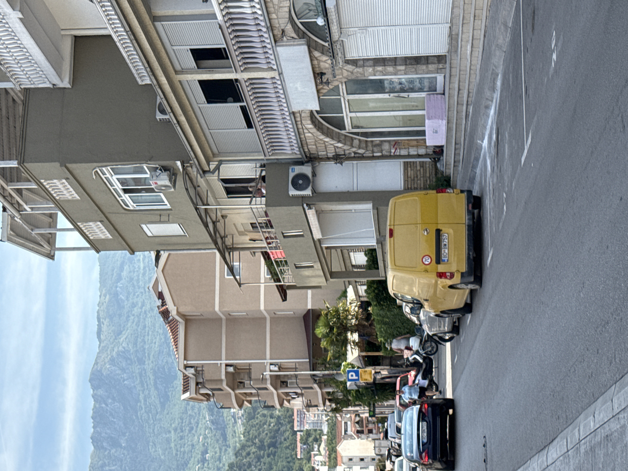 The image depicts a street scene in Budva, Montenegro, with a backdrop of lush green mountains. In the foreground, a yellow van with the license plate "BO BM 757" is parked on the right side of the road, displaying a "70" speed limit sign. To the left of the van, a silver car is also parked. Further down the street, a dark blue sedan with the license plate "BO BM 702" is visible, and a woman in a light blue top and dark shorts is walking on the sidewalk. Several other vehicles are parked or driving on the street.
The buildings lining the street are multi-story structures with balconies and air conditioning units. One building on the right features a stone facade with arched doorways and shops with roll-down shutters. Another building to the left is a beige, multi-story apartment building with numerous balconies. In the background, the verdant mountains rise, with some smaller buildings visible on their slopes. A blue and yellow parking sign is posted on a pole on the left side of the street. A palm tree and other greenery are present between the sidewalk and the buildings. The sky is mostly clear with some scattered clouds. A stack of pink insulation panels is visible on the steps leading up to one of the buildings.