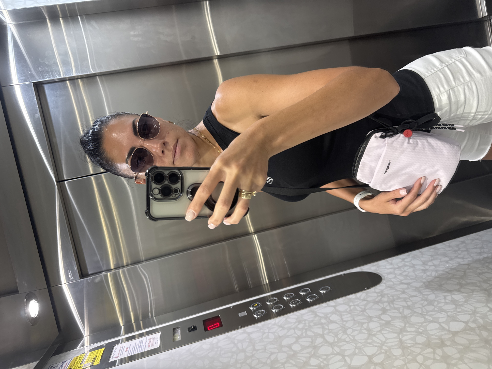 A woman is taking a mirror selfie in an elevator. She has dark hair pulled back and is wearing sunglasses, a black tank top, white shorts, and a small white crossbody bag. She has a gold necklace and two rings on her left hand. The elevator walls are brushed metal, and there are buttons and a display panel on the left side. The woman is holding a smartphone in her right hand, angled to capture the reflection. The floor number "0" is displayed in red. The background of the elevator walls has a subtle textured pattern. The image was taken in an elevator in Netanya, Israel.