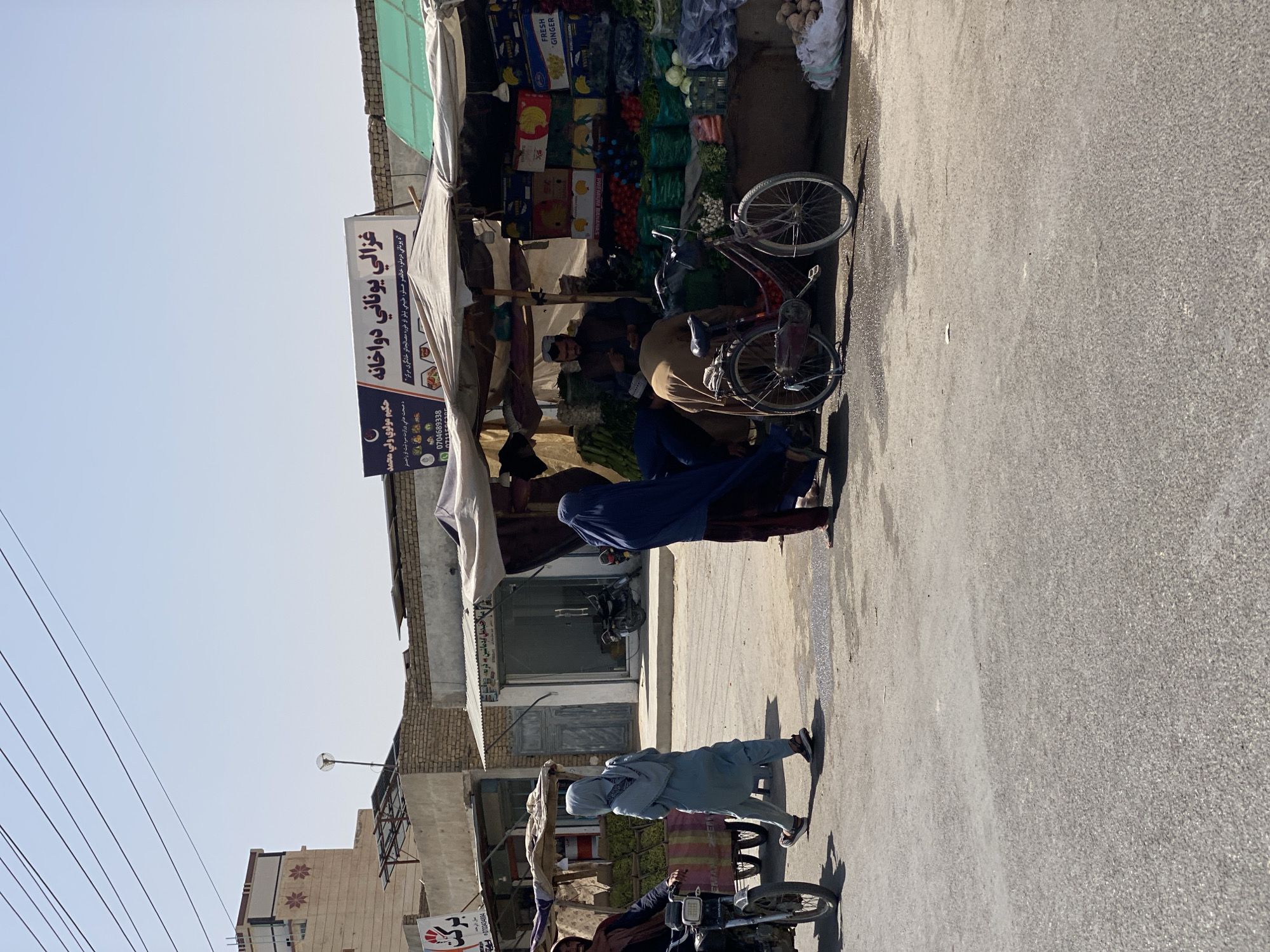 The image depicts a street scene in Kandahar, Afghanistan, with a focus on a market stall selling fresh produce. The stall is covered by a canopy and displays a variety of fruits and vegetables, including what appear to be bananas, tomatoes, cucumbers, and leafy greens. Several boxes of produce are stacked behind the stall.
In the foreground, a red bicycle is parked next to the stall. People are interacting with the market. A person in a dark blue burqa is standing to the right of the stall. A person in a light blue headscarf and traditional clothing is walking away from the stall towards the left. Another person, partially obscured, appears to be bending down near the stall. On the left side of the image, a man is riding a motorcycle, and a woman in a headscarf is walking beside it.
The environment suggests an outdoor marketplace. Buildings with a light-colored facade are visible in the background, along with power lines and a satellite dish. The ground is paved and appears to be a street or a wide walkway. The lighting indicates it is daytime with clear skies. A sign with Arabic script is visible above the market stall.
