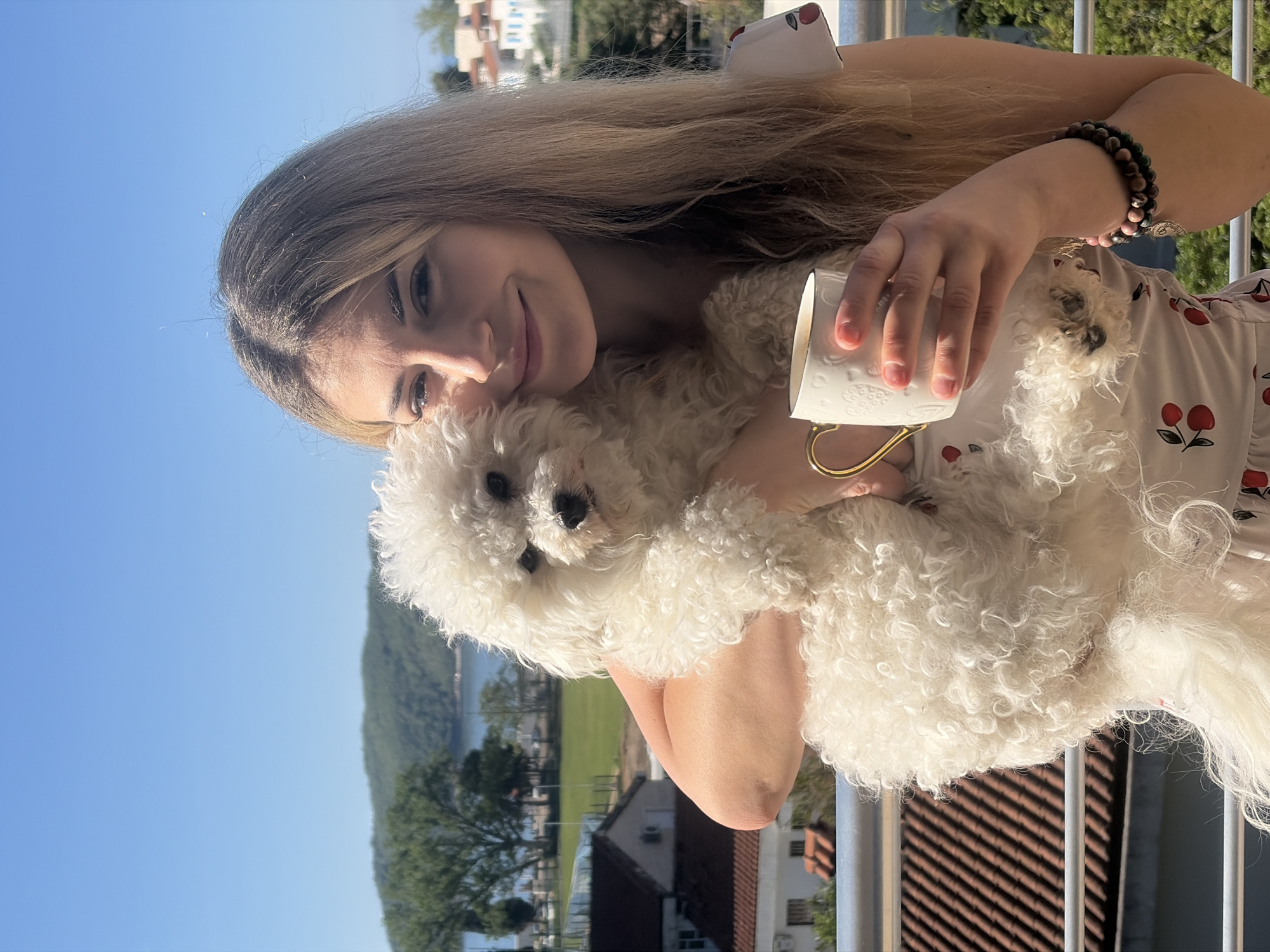 A woman is on a balcony holding a small white Bichon Frise dog and a white ceramic mug with a gold handle. The woman has long blonde hair and is wearing a dress with a cherry pattern. The dog is nestled against her chest. In the background, the scene shows buildings, trees, and the coastline. The location is Budva, Montenegro, based on the characteristic landscape.