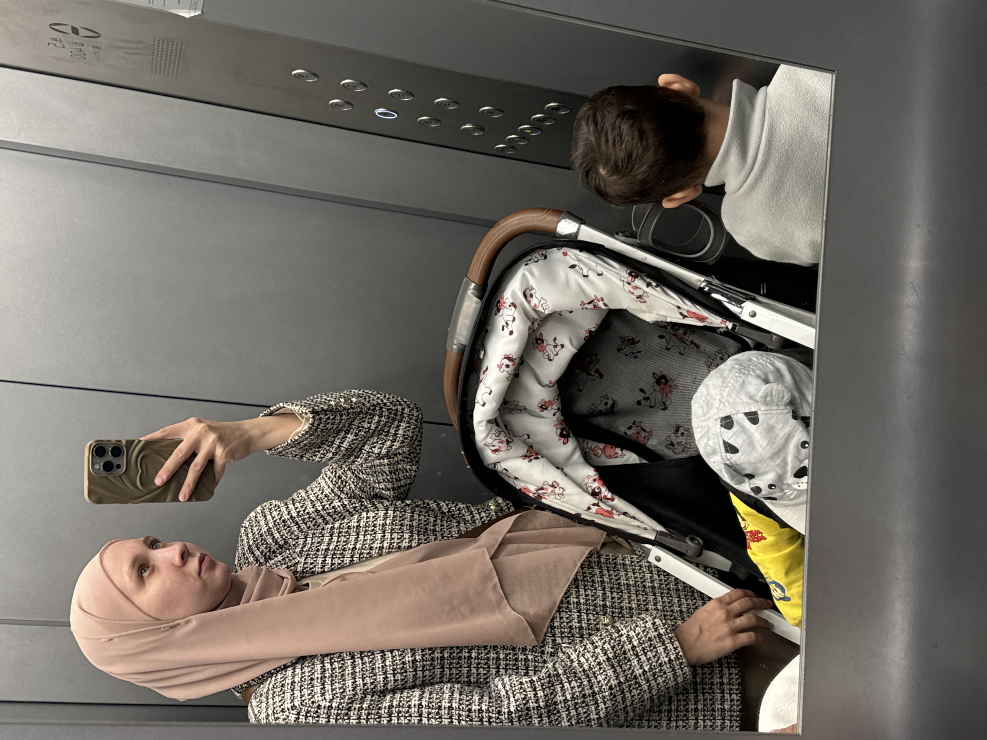 The image depicts a woman in a tweed jacket and a light pink hijab, holding a cell phone and taking a selfie in what appears to be an elevator. A baby stroller with a child inside is also present in the elevator. The interior of the elevator is gray, and there is a panel with elevator buttons visible. The location is unknown and cannot be determined based on the image.