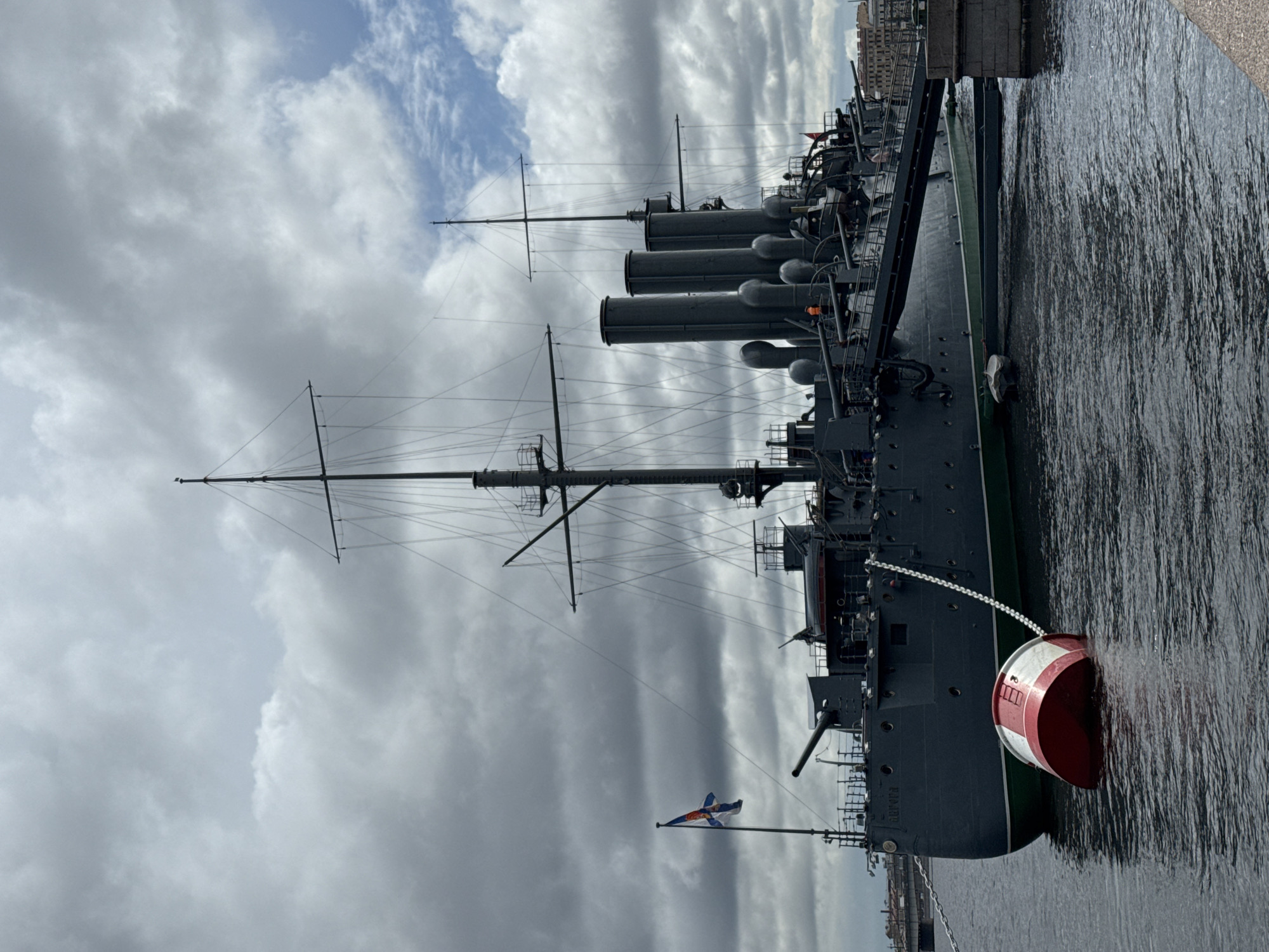 The image shows the cruiser Aurora, a historical warship, docked in Saint Petersburg, Russia. The ship is gray with a tall mast, multiple smokestacks, and cannons. A red and white buoy is attached to the side of the ship. A Russian naval ensign flies from the stern. The sky is cloudy with patches of blue. The ship is in the Neva River.