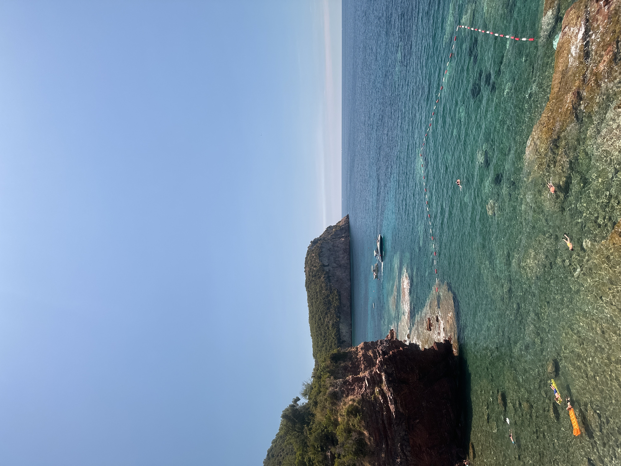 This image depicts a scenic view of the Adriatic Sea near Sveti Stefan, Montenegro, on a clear, sunny day. The foreground features rocky shorelines and shallow, crystal-clear turquoise water. Several individuals are visible enjoying the water; some are floating on inflatable rafts, including an orange one and a yellow one, while others appear to be swimming or snorkeling. A rope with buoys delineates a swimming area in the sea.
In the mid-ground, the water deepens to a rich blue, and two boats are present: a white motor yacht and a darker speed boat, both at anchor. Further in the distance, a large, verdant, rocky headland juts out into the sea. The backdrop is dominated by a vast expanse of clear blue sky. The overall scene is one of a tranquil coastal environment with recreational activities taking place.