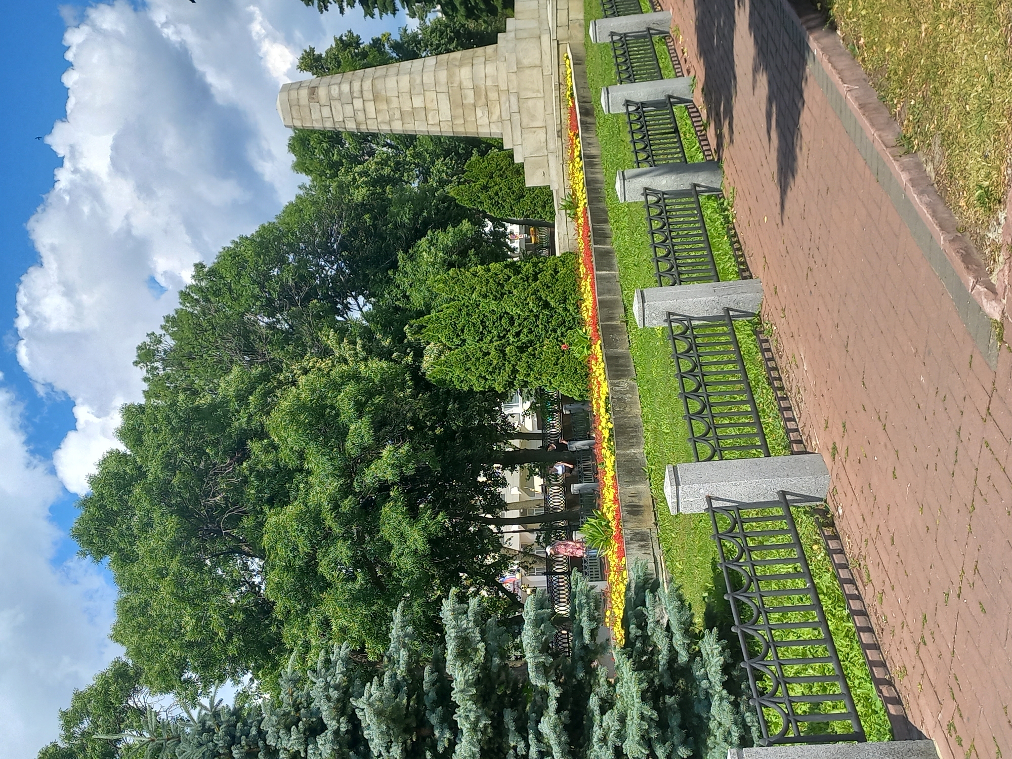 The image depicts a memorial in Victory Park, Ulyanovsk, Russia. A tall, stepped stone monument stands prominently on the right side of the frame, surrounded by manicured greenery and a flowerbed featuring yellow and red blooms. A dark metal fence with decorative elements separates the park's lawn from a brick-paved walkway. A large blue spruce tree is visible on the left, with lush green deciduous trees forming a backdrop. In the background, beyond the fence, a few people are visible, with buildings and more trees further in the distance. The sky above is a bright blue with scattered white clouds.