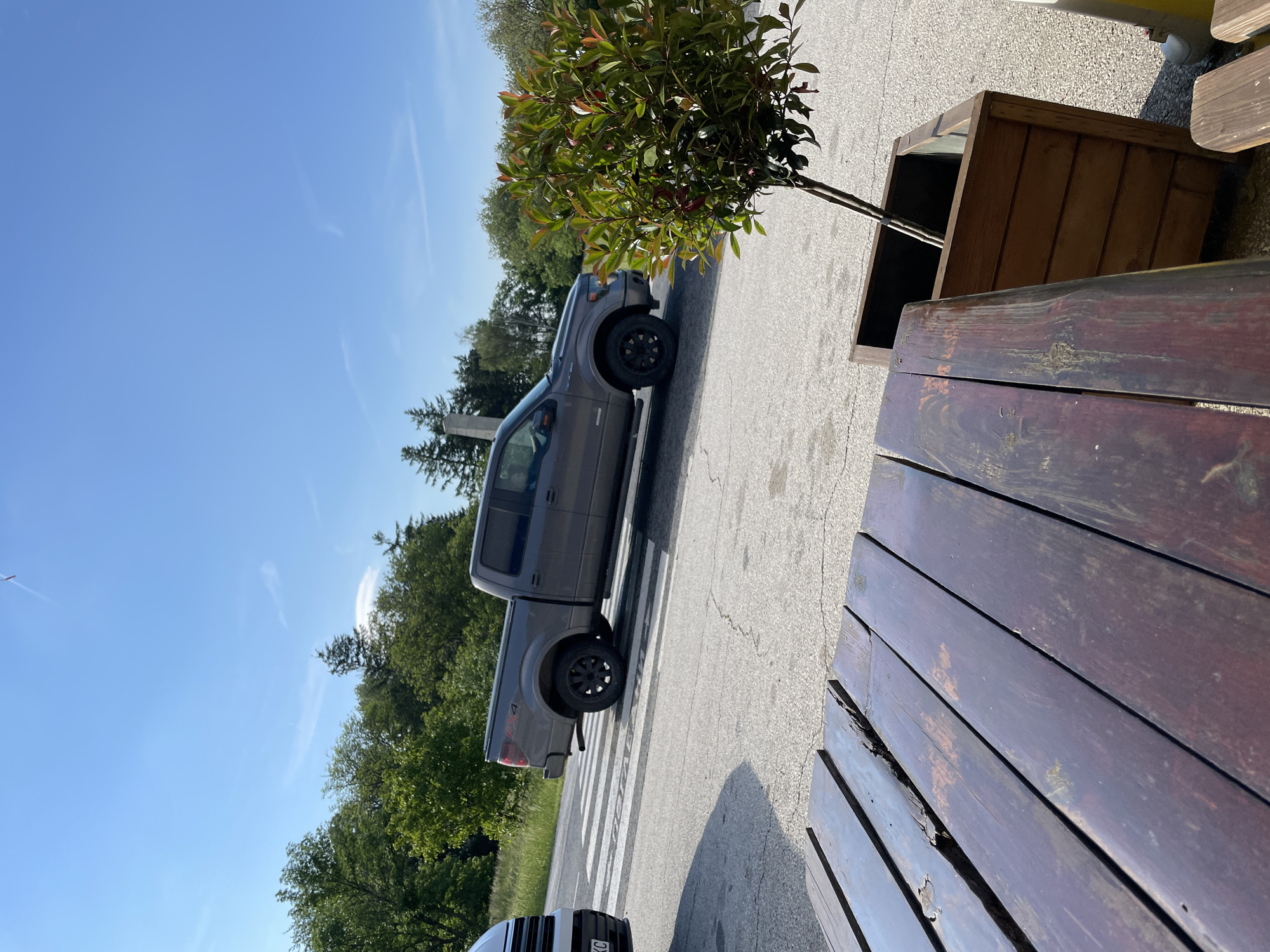 The image depicts an outdoor scene in Sheynovo, Bulgaria. A dark-colored pickup truck is parked on a zebra-striped crosswalk. A wooden bench sits in the foreground, with a potted tree to its right. In the background are various trees and a clear blue sky. A portion of another vehicle is seen at the very bottom of the image. There are no people visible.