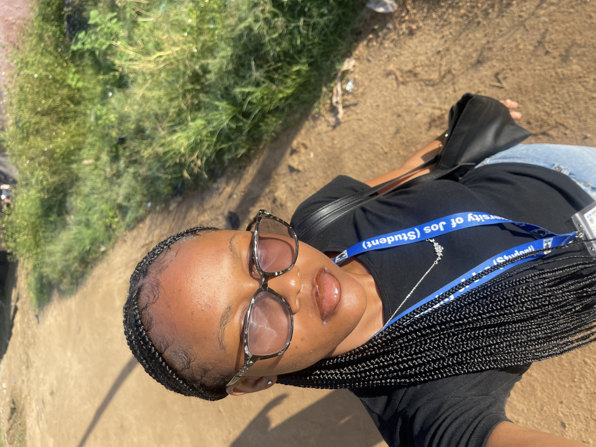 A young Black female stands on a dirt path, captured from a high-angle perspective. She has her hair braided in thin cornrows and wears large, patterned glasses. Her lips are shiny with lip gloss. Around her neck, a blue lanyard with text, possibly indicating student identification from the University of Jos, is visible, along with a delicate necklace. She is dressed in a black t-shirt and light-wash jeans. A black handbag hangs from her left shoulder. The background consists of a dirt path and overgrown green vegetation, suggesting an outdoor, possibly rural or semi-urban setting. The lighting indicates it is daytime, with strong sunlight casting shadows. The location is identified as Rafin Par, Nigeria.