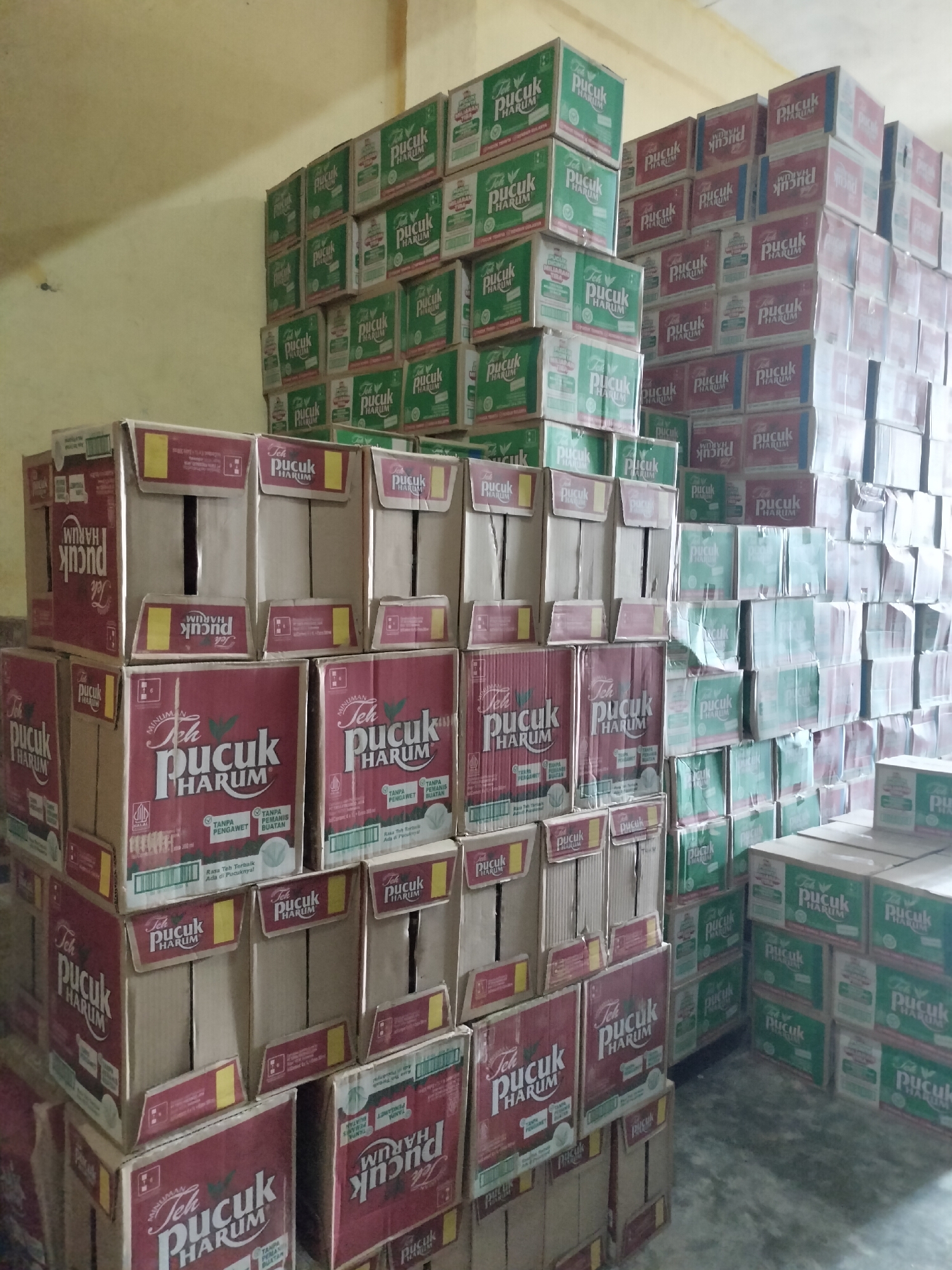 The image depicts a storage room filled with stacks of packaged tea boxes. The boxes are predominantly red and green and feature the brand name "Teh Pucuk Harum". The arrangement suggests a commercial setting, possibly a warehouse or a stock room for a retail business. The lighting is dim, casting shadows on the stacked boxes. The background shows a plain, lightly colored wall, and the floor appears to be concrete. The overall scene is static and focuses solely on the inventory of tea products. The presence of the "Teh Pucuk Harum" brand, which is a popular Indonesian tea, strongly suggests the location is within Indonesia. While the image does not contain any direct visual cues indicating Langsa, Indonesia specifically, this brand's prevalence in the Indonesian market makes it a plausible location.