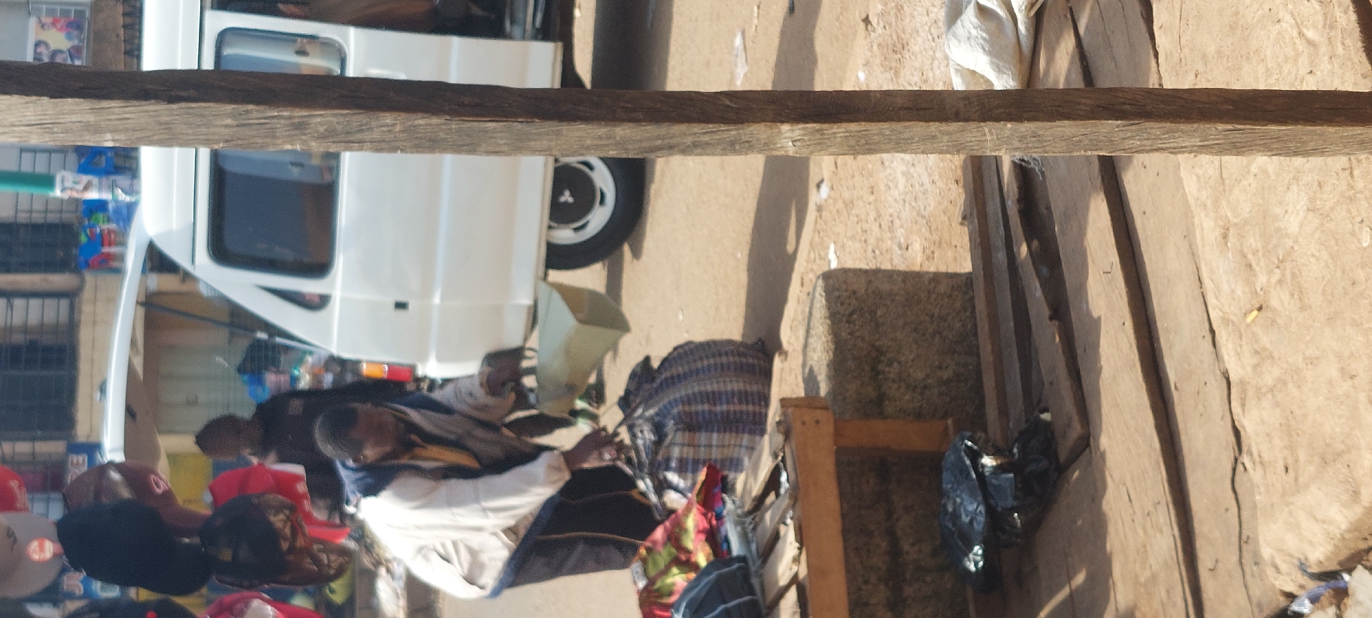The image depicts an outdoor scene, likely a market, in Bukuru, Nigeria. A man, dressed in a light-colored jacket and dark pants, is seen reaching down to pick up a large, checkered duffel bag. He is also holding a tan-colored, rectangular container in his right hand. In the background, a white van with its rear doors open is parked. Various items are visible, including what appear to be bags of goods, clothing items on display, and what looks like a wooden stall or table in the foreground. The ground is unpaved, with a dirt and gravel surface. A prominent wooden post stands vertically in the right side of the frame. The overall impression is of a busy, informal marketplace.