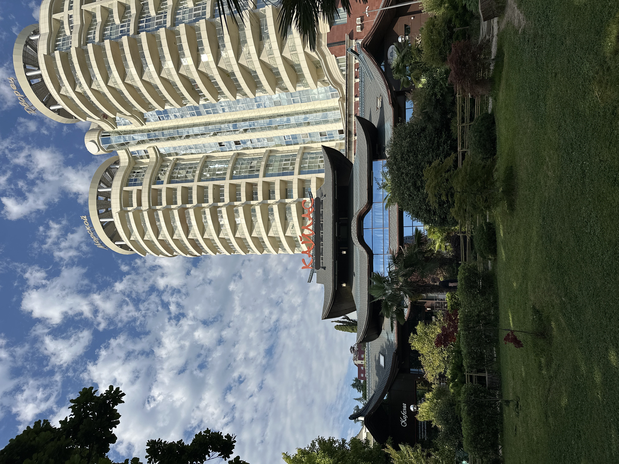 The image depicts the Kagemu restaurant and the nearby high-rise residential buildings "Morskoy" and "Dvoretz" in Sochi, Russia. The Kagemu restaurant, with its distinct traditional East Asian roof architecture, is situated in the foreground, surrounded by lush greenery and trees. The residential buildings, characterized by their modern, curved design and numerous balconies, tower over the restaurant in the background. The sky is blue with scattered white clouds. A person is visible walking on the right side of the restaurant complex. The scene is set in a well-maintained park-like environment.