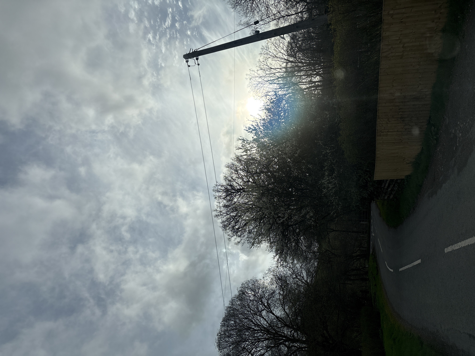 The image depicts a rural road scene in Shrewsbury, United Kingdom. An asphalt road curves slightly to the right, marked with a broken white line. The road is flanked by a dense treeline on the left, partially obscuring the sun and sky, with bare branches indicating the season is likely late autumn or winter. A wooden fence runs along the right side of the road, bordering a grassy area. An electrical pole with power lines extends diagonally across the frame, contrasting with the cloudy sky. The sky is filled with layers of grey and white clouds. No people or animals are visible.