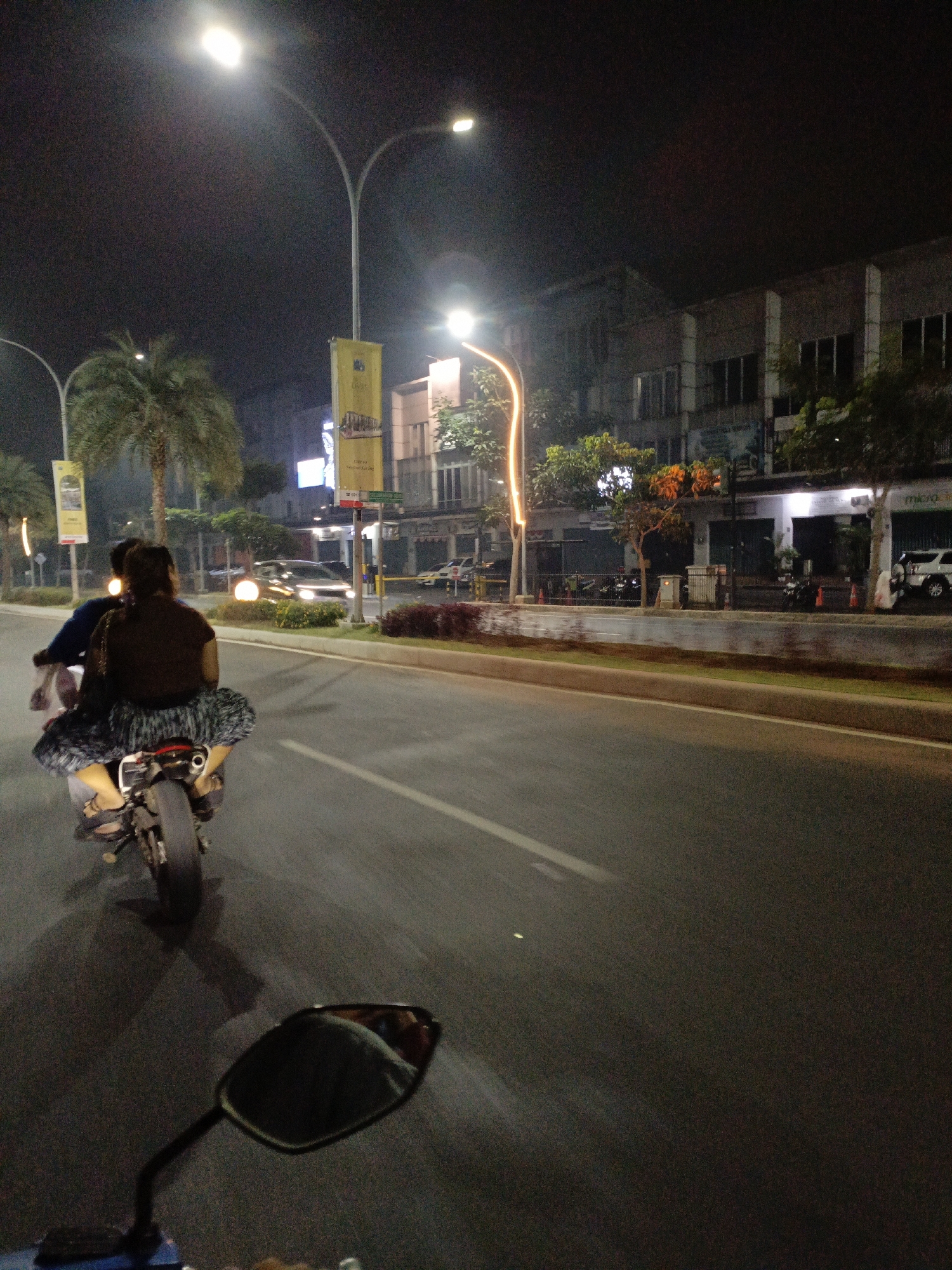 This image captures a nighttime scene on a city street in Lambangjaya, Indonesia. The perspective is from the viewpoint of someone riding a motorcycle, looking forward.
In the foreground, the motorcycle's side mirror is visible, reflecting a person wearing a grey top. The rear of the motorcycle is also in view, with its wheel and exhaust pipe clearly discernible.
On the motorcycle, a person is riding as the pillion passenger, wearing a dark brown top and a patterned skirt. Their legs are visible, wearing sandals. They are seated behind the rider, whose torso is partially visible.
The street itself is paved, with a lane divider line visible. The background depicts a city environment at night. Lampposts with bright lights illuminate the scene. Adjacent to the street is a sidewalk with a concrete barrier. Beyond the sidewalk are buildings, characterized by their windows and architectural features. Some of these buildings appear to be shops or commercial establishments, with illuminated signs and windows.
Trees, including palm trees, are scattered along the sidewalk, their foliage visible against the night sky. There are also some orange traffic cones on the sidewalk. Several vehicles, including cars and what appear to be motorcycles parked along the buildings, are present in the background. The overall atmosphere is one of urban nighttime activity.