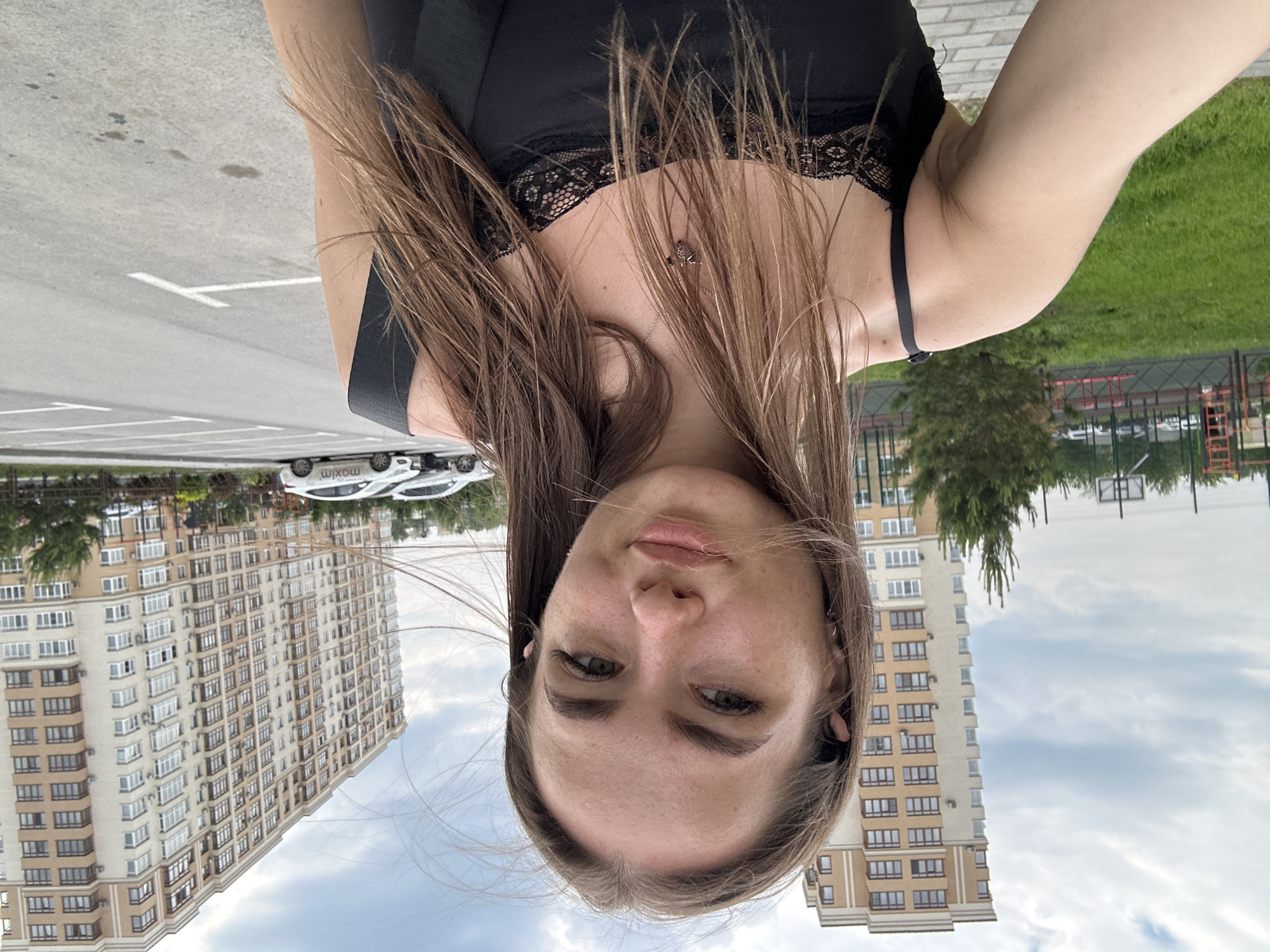 A young woman with long brown hair is taking a selfie outdoors in a paved area. She is wearing a black tank top with lace trim. Behind her, there are two large, modern apartment buildings, a grassy area with a playground, and a parking lot with several cars. The sky is cloudy.