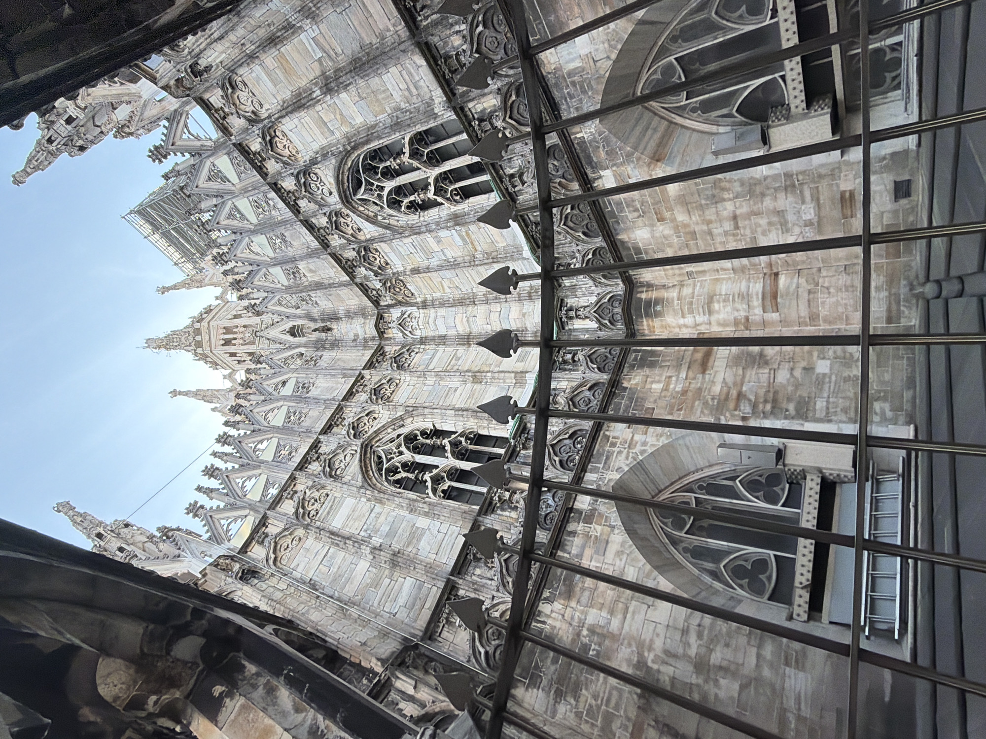 The image captures a low-angle view of the Milan Cathedral (Duomo di Milano) in Milan, Italy. The cathedral's facade, adorned with Gothic architecture, is visible, with its spires, windows, and intricate details. A metal grid or fence-like structure is in the foreground. The sky is pale blue, suggesting daylight. No people are visible.