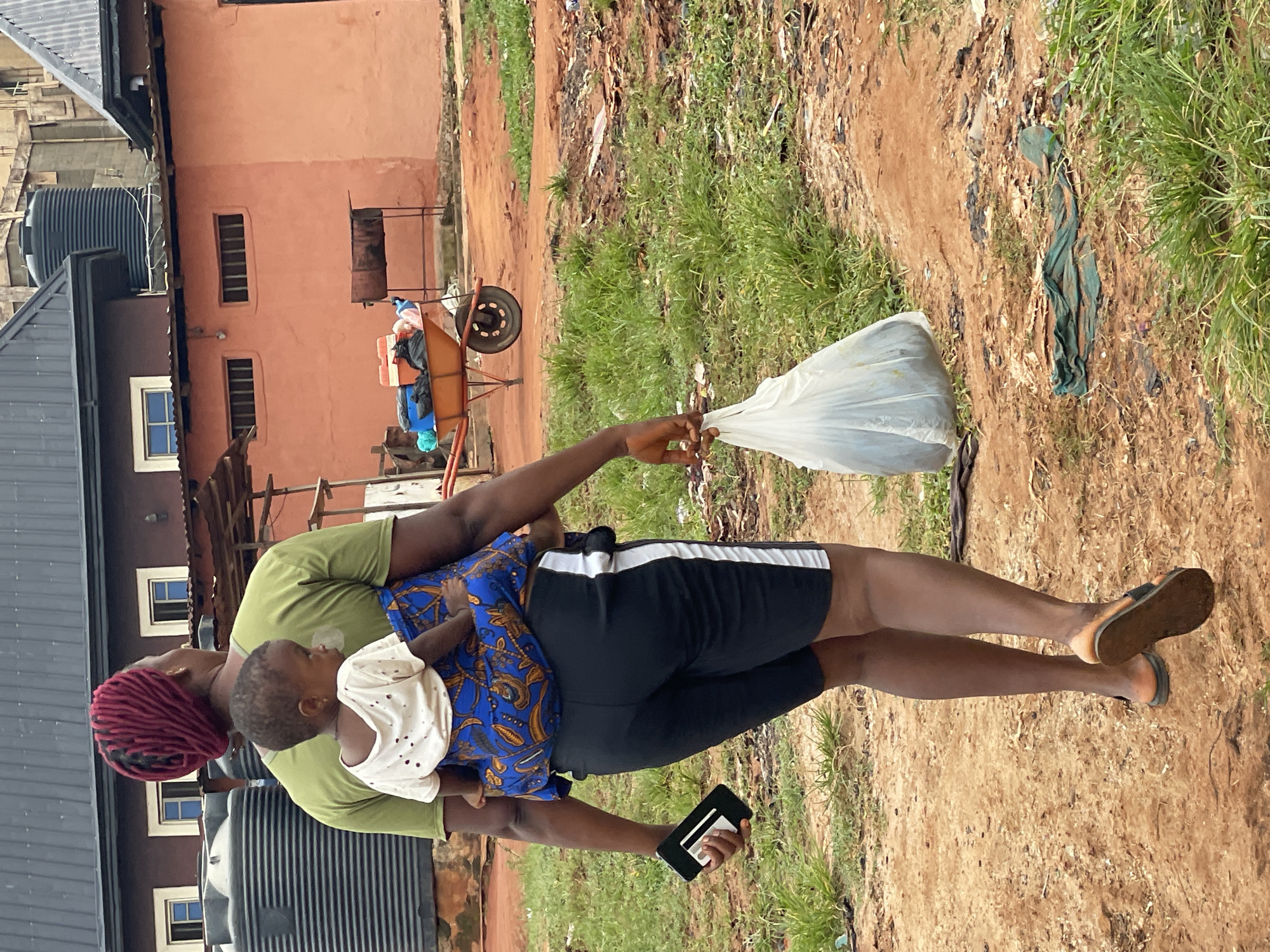 A woman is carrying a child, holding a plastic bag, and a mobile phone outdoors in Nkpor, Nigeria. The woman wears an olive green shirt, black shorts with a white stripe, and brown sandals. She has red braids. The child wears a white shirt and a blue and brown patterned dress. The woman is holding a clear plastic bag in her right hand and a black mobile phone in her left hand. The background includes a dirt path, grass, scattered debris, a rusty wheelbarrow, a red building, a black water tank, and other buildings.