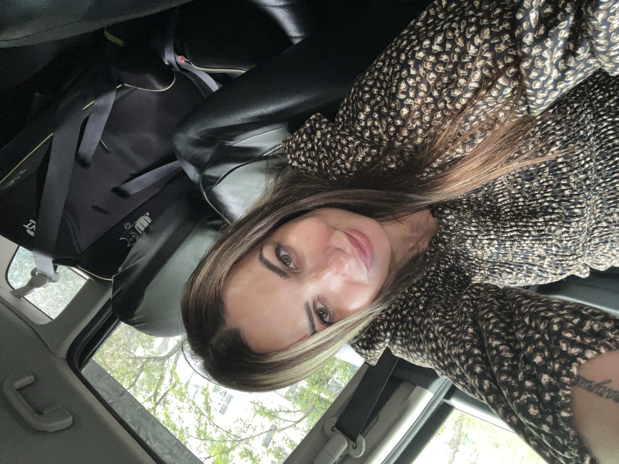 A woman with brown hair and brown eyes is taking a selfie in the backseat of a car. She is wearing a brown and black patterned dress. The car appears to be parked. Trees and buildings are visible through the windows. There is a car seat behind her.