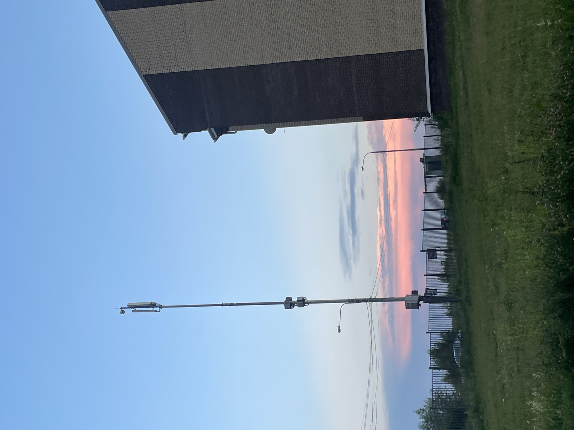 The image shows a cell tower in Zverevo, Russia, next to a brick building. The sky is a pale blue with some pink and purple clouds. In the foreground, there is a patch of green grass with some wildflowers. A black metal fence runs across the middle of the image, and behind it, there are some cars and trees. A street lamp is visible next to the cell tower. The building appears to be modern, with dark and light brick sections. The cell tower is tall and slender, with several antennas attached to it. The overall scene suggests an urban or suburban environment at dusk.