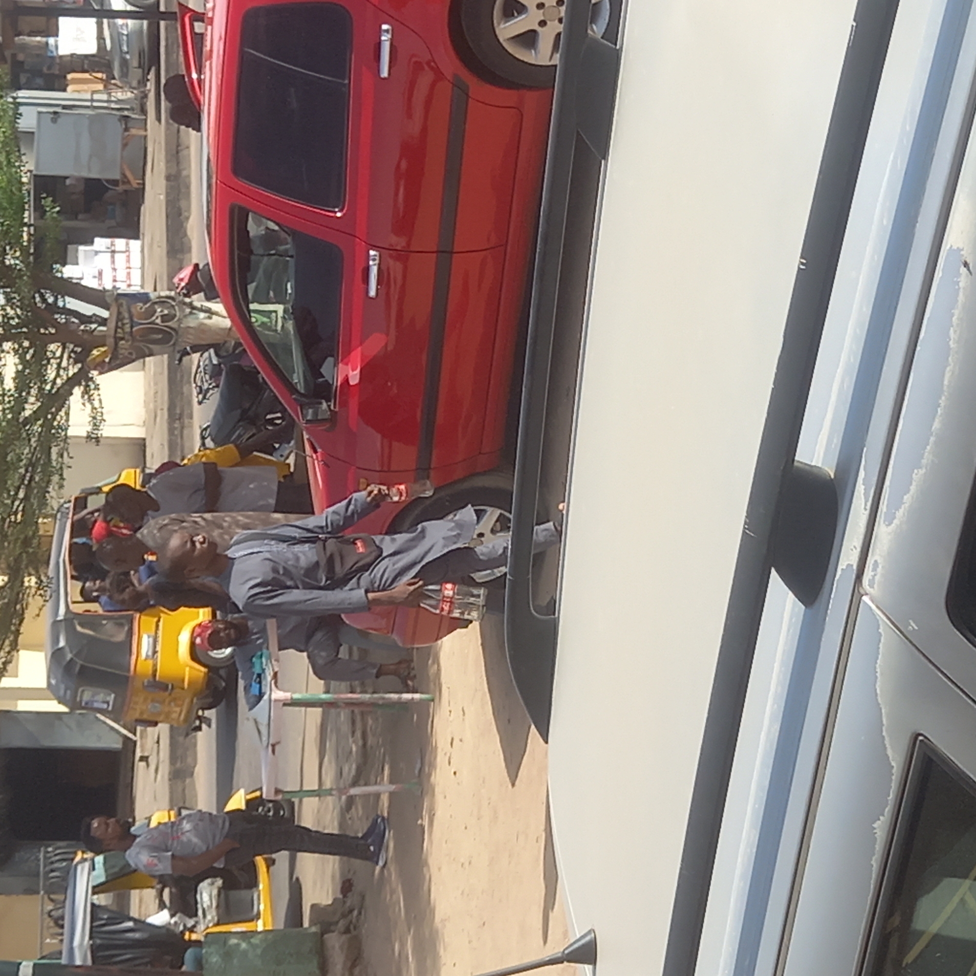 The image shows a street scene in Dadi Riba, Nigeria. A red SUV is parked next to a larger white vehicle. Several people are standing and sitting nearby, including a man in a gray outfit holding two bottles of Coca-Cola and walking adjacent to the red SUV's rear wheel. A yellow and black auto rickshaw ("keke") is visible with multiple passengers. The scene also includes what appears to be street vendors and other bystanders.