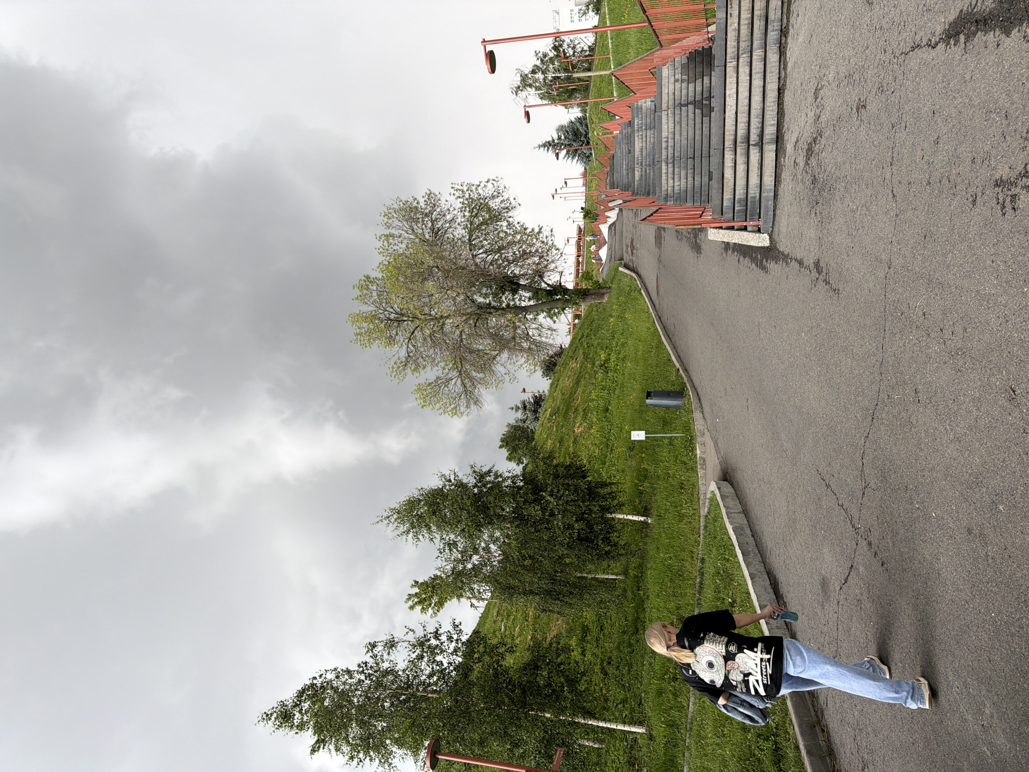 The image depicts a view of a park pathway and an outdoor staircase in Nizhny Novgorod, Russia. A young woman with blonde hair tied in a ponytail, wearing a black t-shirt with a graphic print and light blue jeans, walks away from the camera on the pathway. She is carrying a denim jacket over her shoulder and holding a blue water bottle. The pathway curves to the right, leading towards a wide stone staircase with red railings, ascending a grassy hill. Trees and green grass are visible on the slopes of the hill. The sky is overcast with dramatic, grey clouds. Streetlights with red posts are visible along the pathway and next to the stairs. The environment appears to be a public park or recreational area, likely designed for walking and climbing stairs.