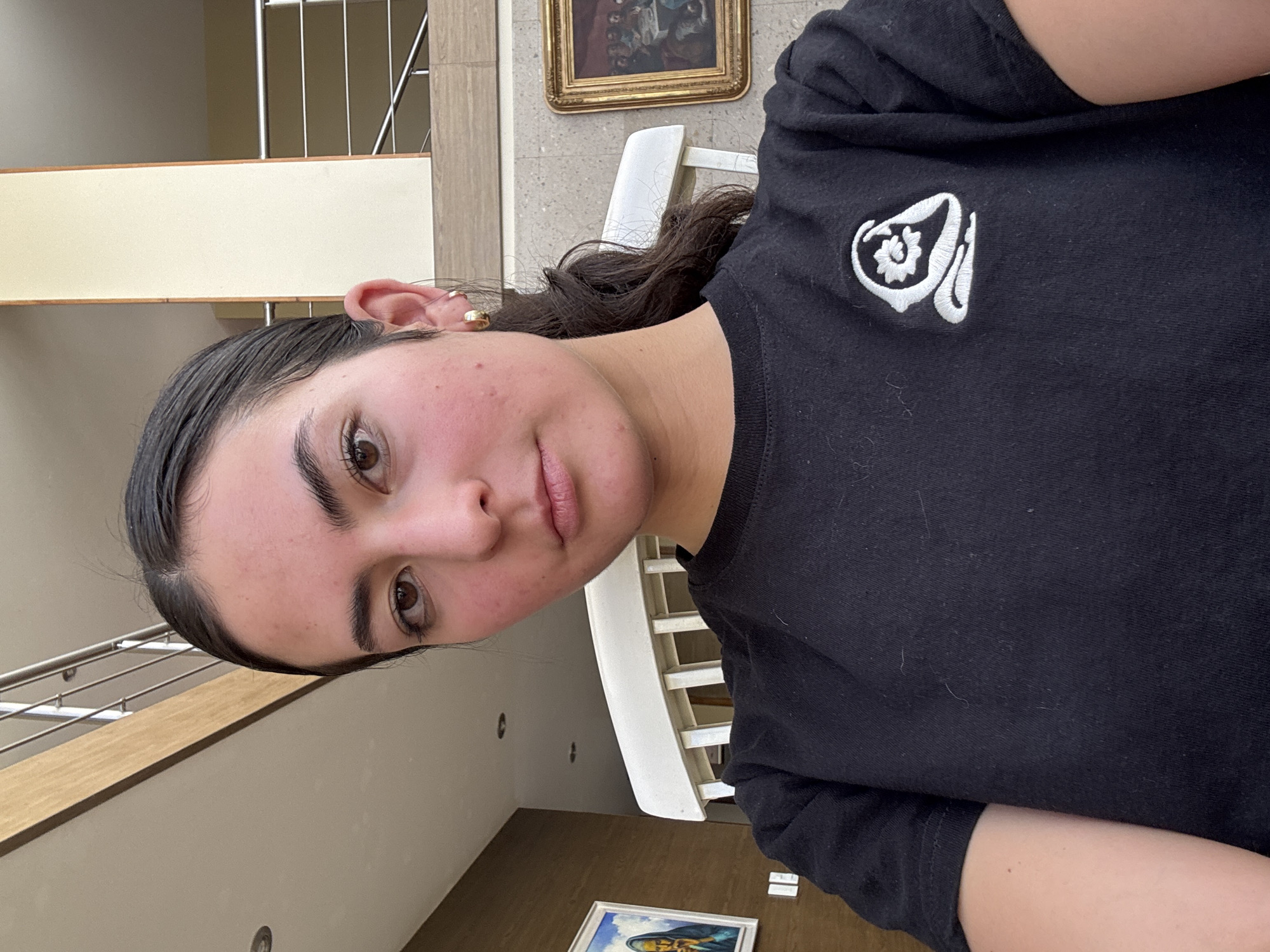 A young woman is depicted in a medium close-up, rotated portrait, facing directly towards the camera with a neutral expression. Her dark hair is pulled back, appearing somewhat slicked or wet. She has dark eyebrows, brown eyes, and wears a small gold hoop earring in her visible left ear. Her fair skin shows some redness on her cheeks and forehead. She is wearing a black short-sleeved t-shirt featuring a white embroidered logo on the upper right chest, which consists of a stylized flower or gear design enclosed within an abstract, fluid shape.
The background suggests an indoor, multi-level architectural space, consistent with a home or building in San Bernardino Tlaxcalancingo, Mexico. To the left, a modern metal railing with horizontal bars and a light-colored wooden handrail is visible. Behind her, a white chair with vertical slats is partially seen. The walls are light-toned, with some areas exhibiting a textured finish. Two framed artworks adorn the walls: an ornate gold-framed traditional painting is visible higher up behind her head, and another, partially visible at the bottom, features a blue and brown abstract artwork. The scene is illuminated by bright, even, natural light, indicating it is daytime. The image itself is rotated 90 degrees counter-clockwise. No discernible text is visible in the frame.
