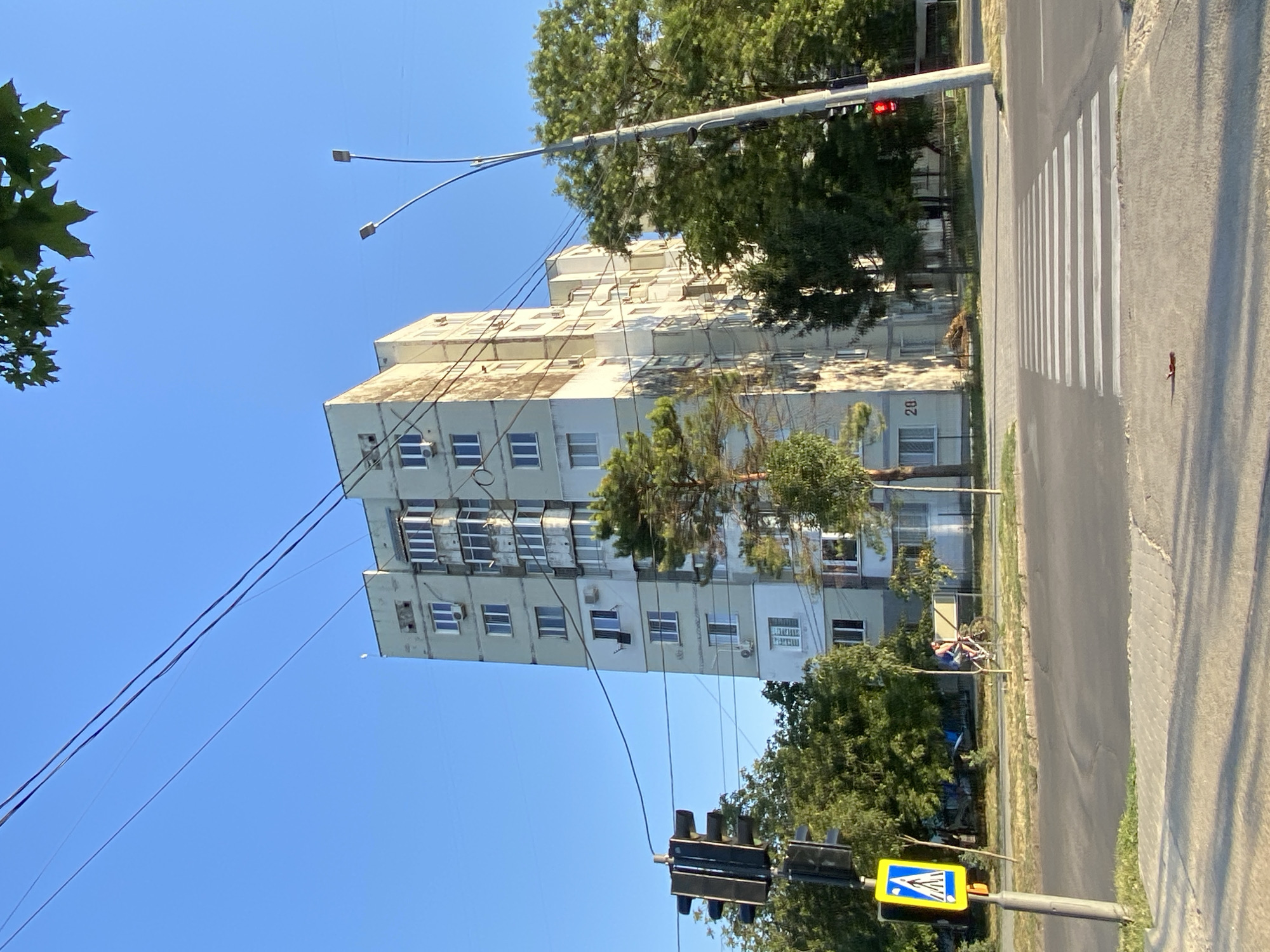A street scene in Chișinău, Moldova, features a tall, light-colored apartment building as the central subject. The building has multiple stories with uniformly spaced windows and air conditioning units. A traffic light with a red light illuminated is visible on the right side of the image. In the foreground, a pedestrian crossing is marked with white stripes on the asphalt. A person is riding a bicycle on the sidewalk to the left, near a pedestrian crossing sign. Several trees with green foliage are interspersed between the building and the street. The sky is clear and blue, indicating a sunny day. Overhead power lines stretch across the frame. The building is marked with the number "20".