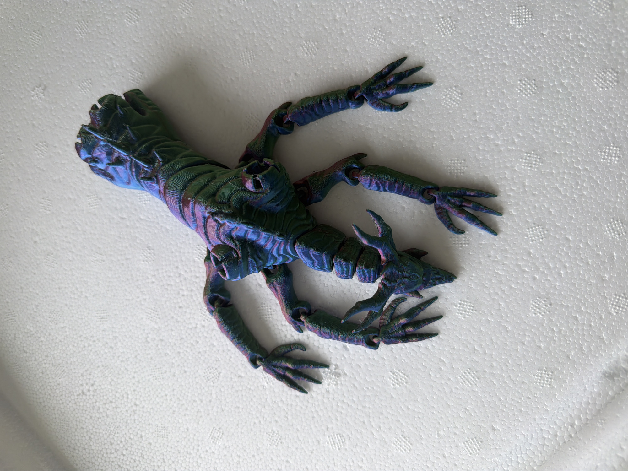A high-angle, overhead shot depicts a single, multi-limbed, articulated creature model positioned on a textured white surface. The creature, approximately 30-40 cm in length, exhibits an iridescent color palette, predominantly shifting between blues, greens, purples, and pinks, typical of multi-color 3D printing filament. Distinct horizontal layer lines are visible across its entire surface, indicating its 3D-printed origin. Its body is segmented and elongated, with a pronounced, textured back and a head featuring two prominent eye-like structures and several pointed, horn-like protrusions. Six articulated limbs extend from its torso, each ending in a five-fingered, clawed hand-like appendage. The creature is oriented with its head towards the bottom-center of the frame and its segmented posterior extending towards the upper-right. It lies on its back, with limbs splayed outwards.
The underlying surface is uniformly white and consists of densely packed, small, spherical pellets, characteristic of expanded polystyrene foam. Several faint, repetitive square patterns, formed by impressed grid-like indentations, are visible across the white surface, particularly in the upper-left, mid-right, and bottom-right sections of the frame. The lighting is bright and even, suggesting an overhead light source, casting minimal discernible shadows.
Location: Unknown, Unknown.
