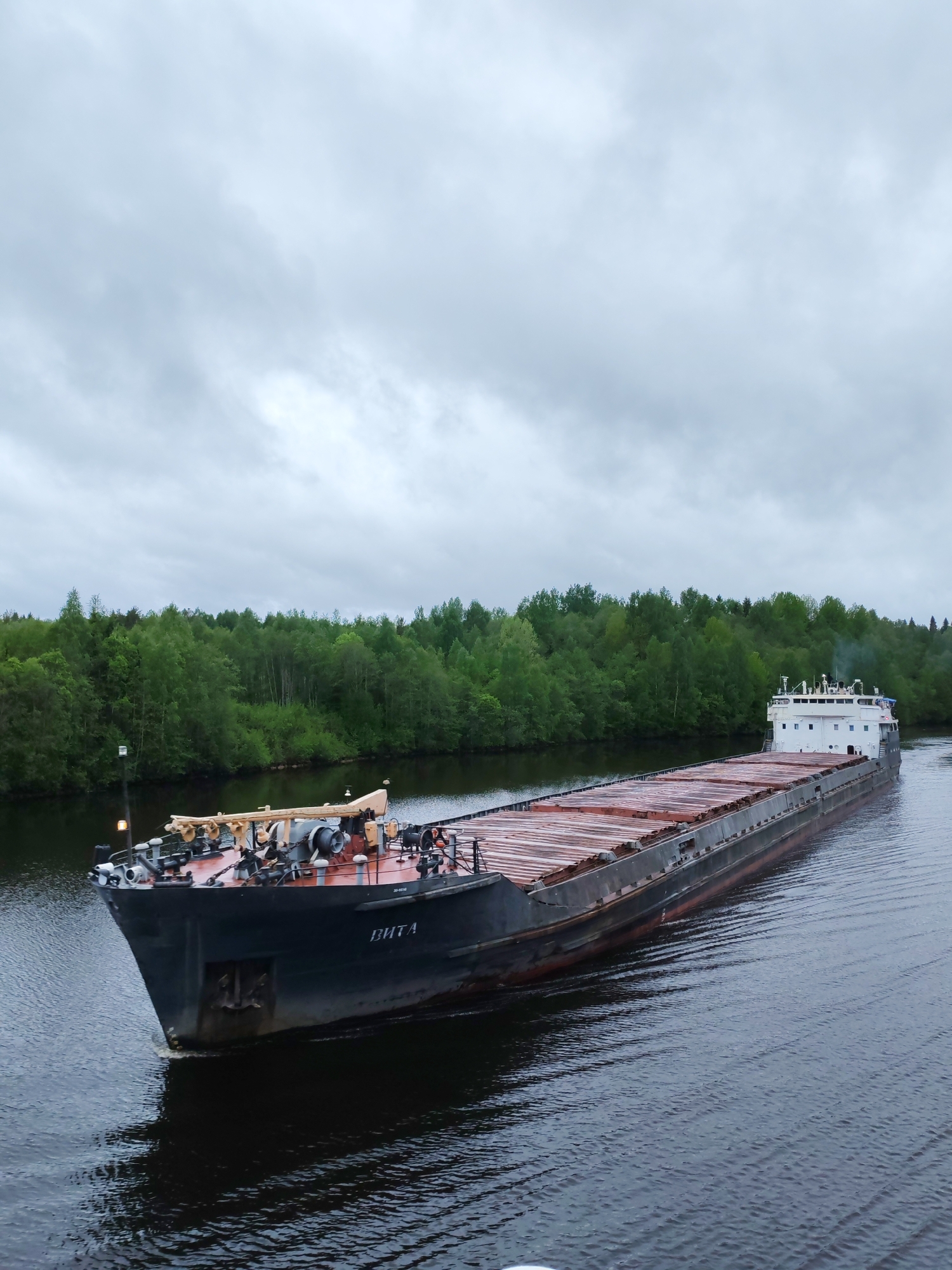 A barge named "ВИТА" is traveling on a river in Depo, Russia. The barge is loaded with what appears to be a cargo of covered materials. Lush green trees line the riverbank. The sky is overcast, suggesting cloudy weather.